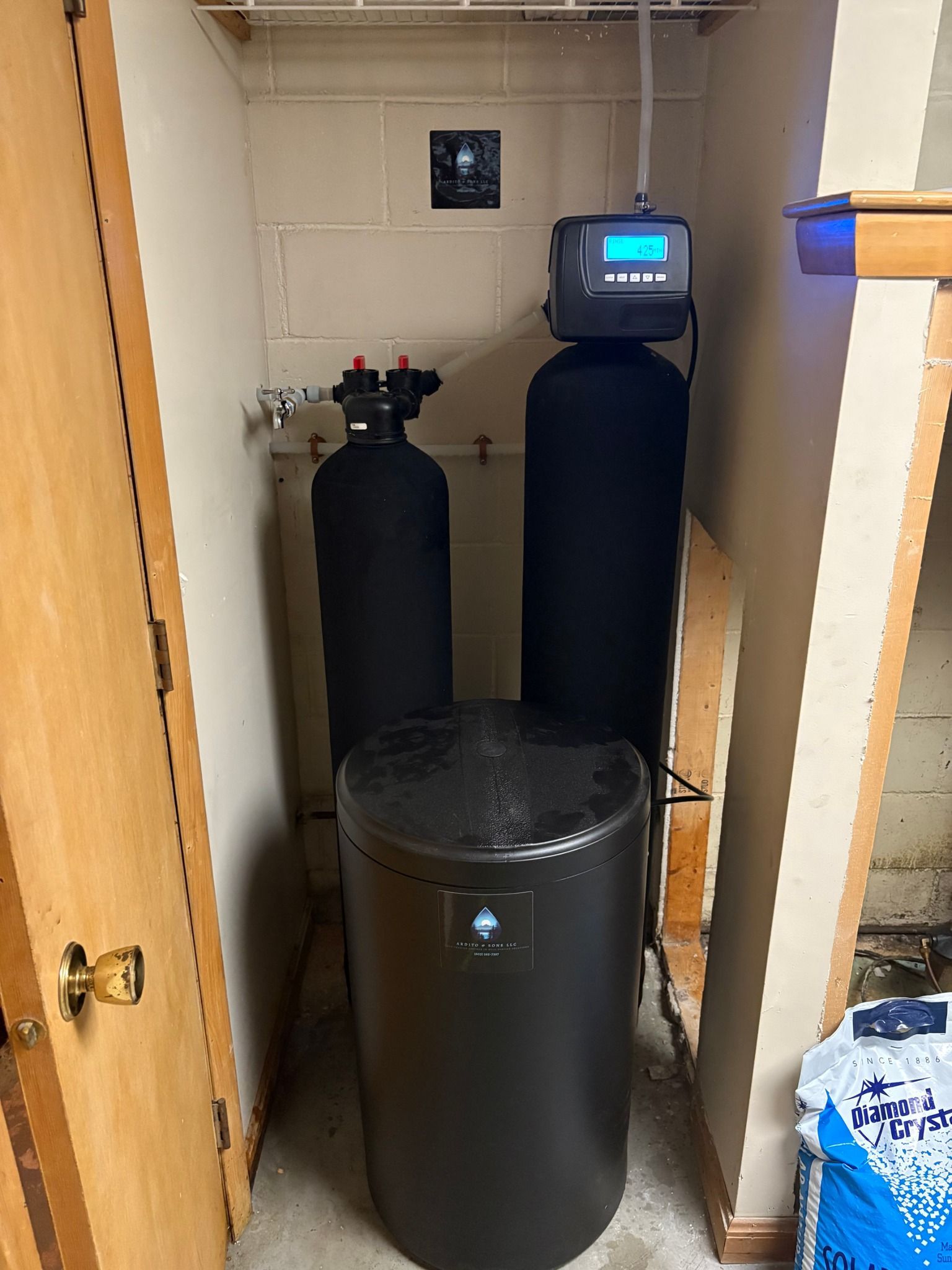 Water softener system in a small utility closet. Black tanks, digital control panel, and salt storage.