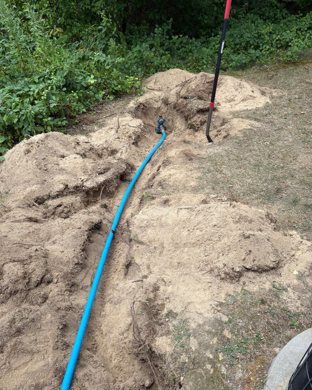Blue pipe in a trench, surrounded by sand, with a shovel and greenery in the background.
