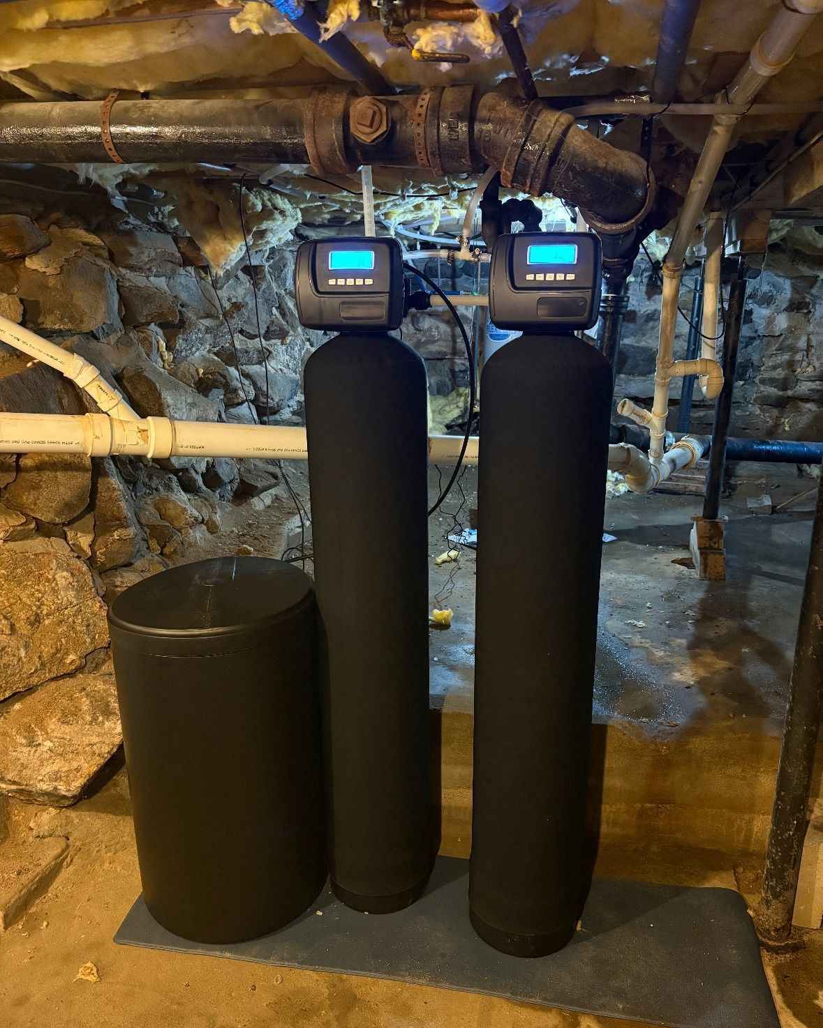 Water treatment system in a dimly lit basement: two tall black tanks with digital displays, and a salt tank.