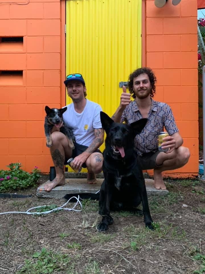 Two Men Crouch With Two Dogs in Front of a Bright Orange and Yellow Building — Eagle Tamer Barbershop in Stratford, QLD