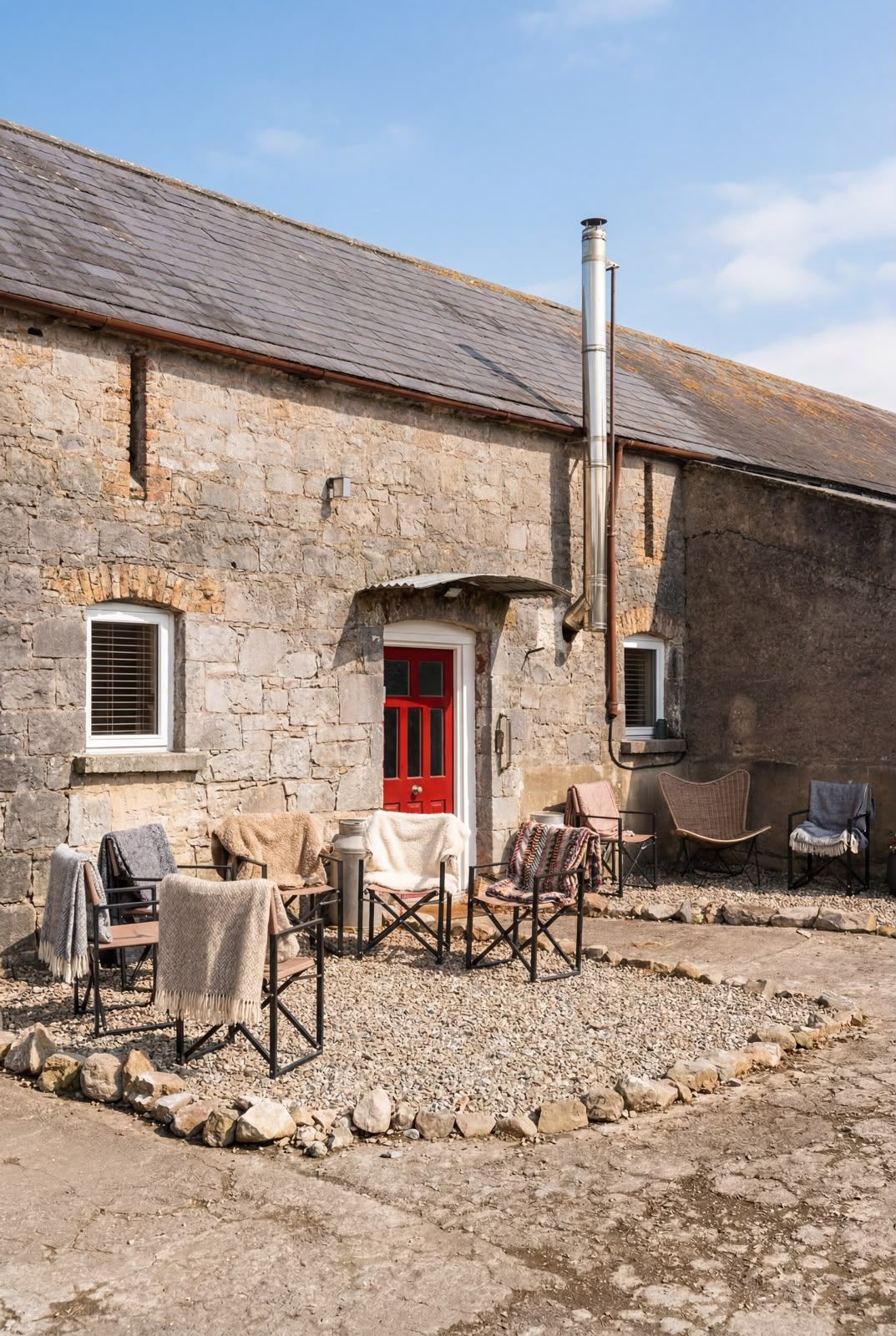 Rustic stone cottage with a red door, small patio chairs, and a chimney under a blue sky.