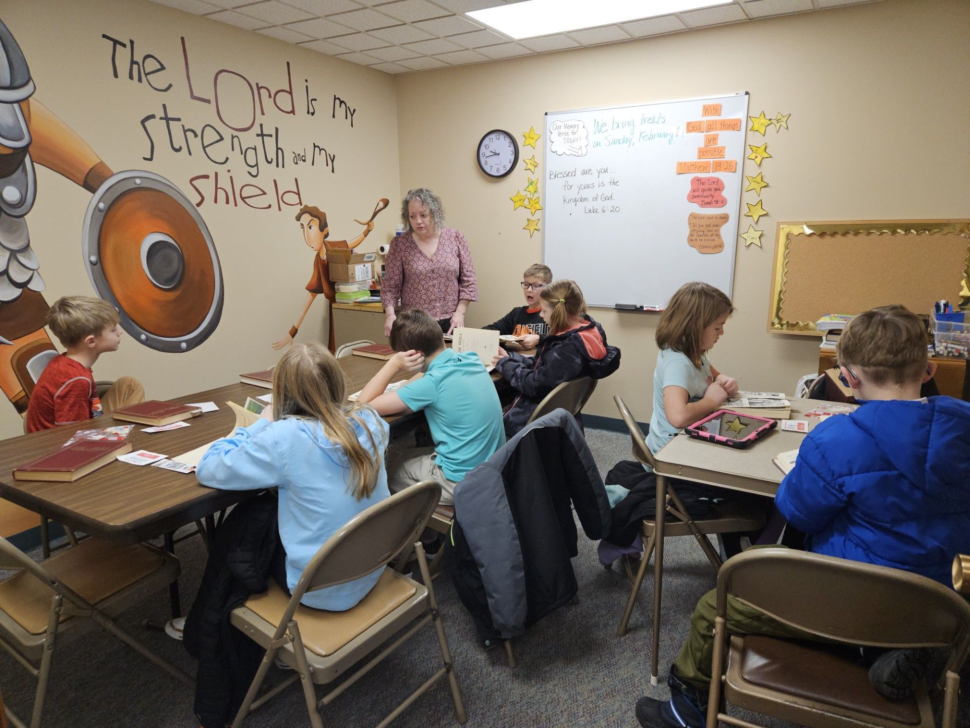 Classroom scene with students at tables and a teacher near a whiteboard and mural at the front.