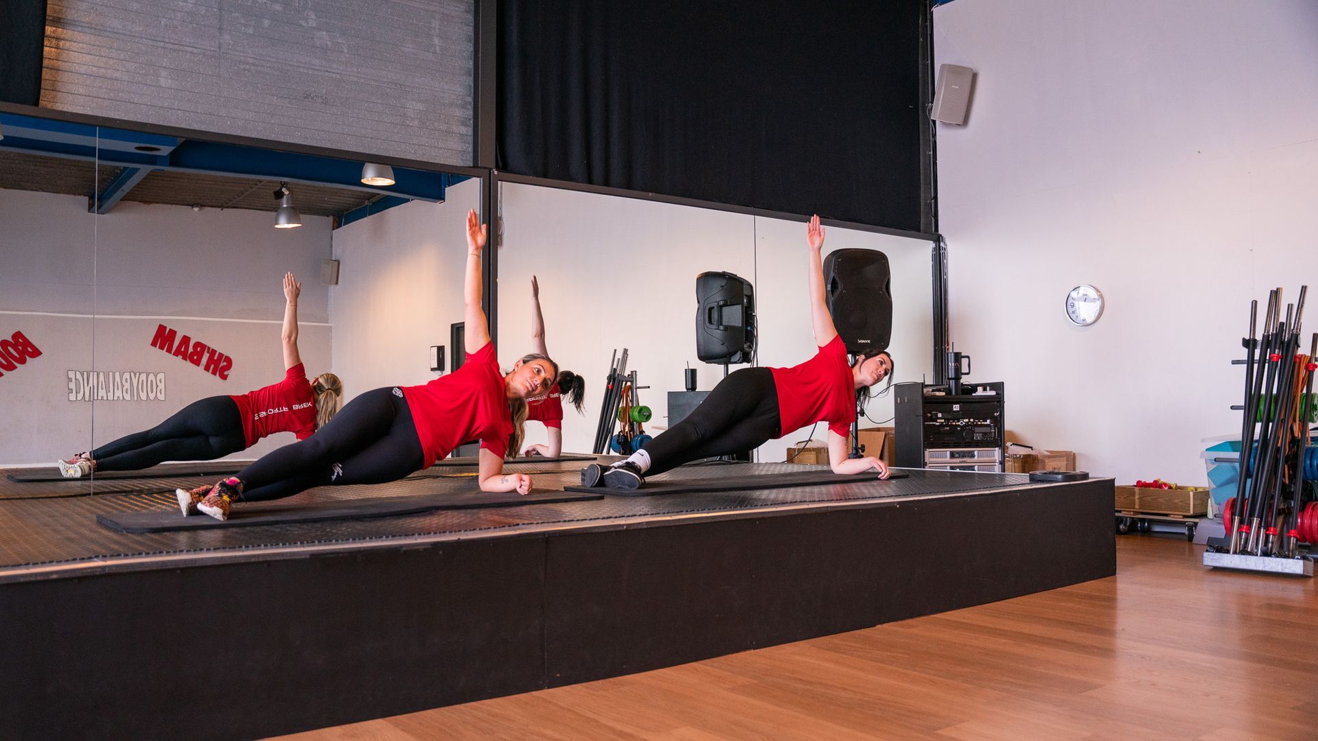 Three people in red shirts do side planks on a stage in a gym.