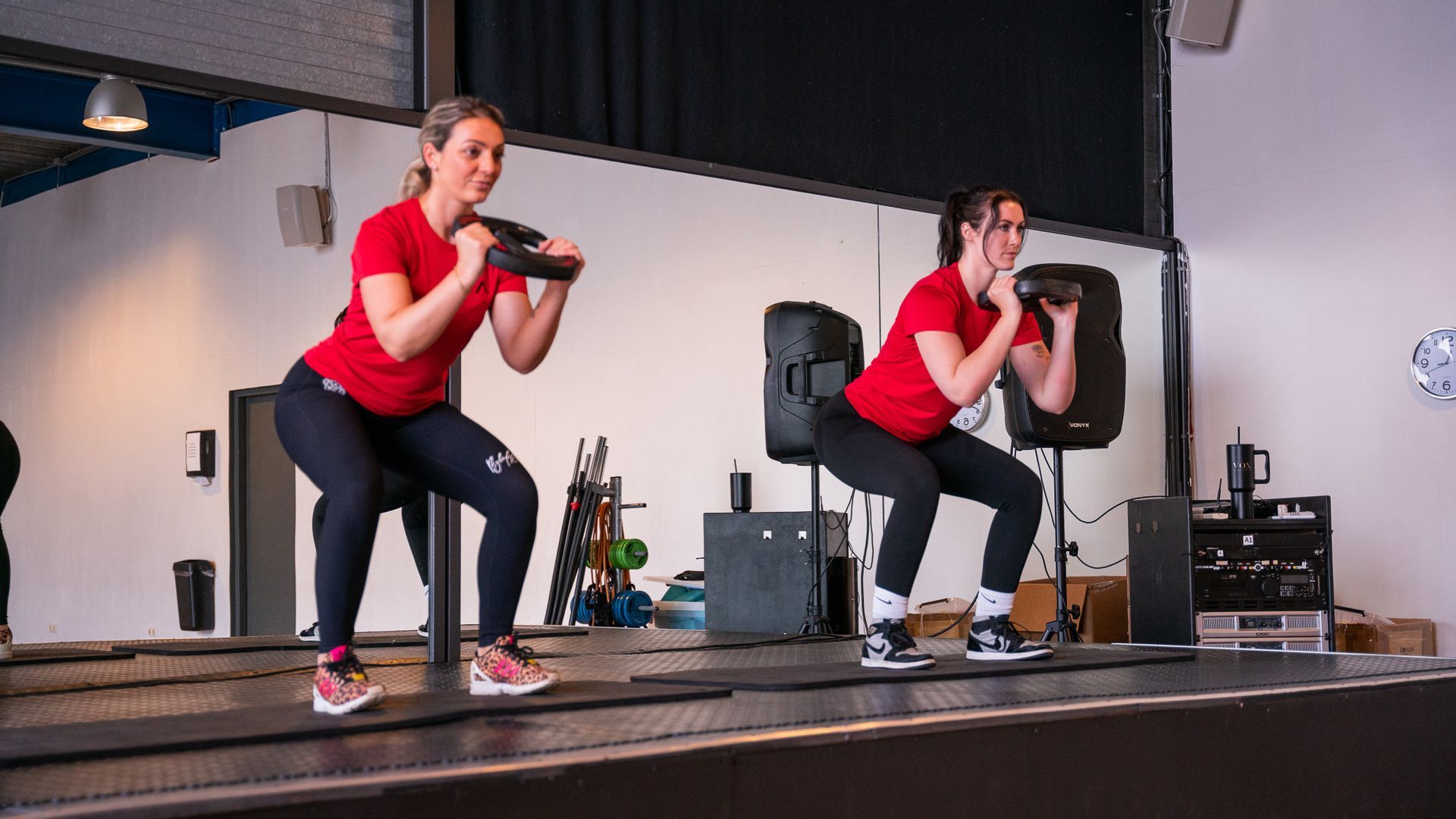 Twee vrouwen doen squats op een podium in een sportschool.