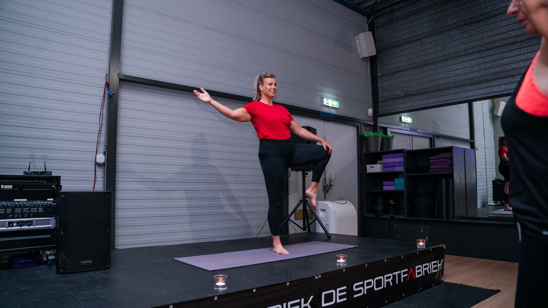 Woman in red top poses on a stage with one leg raised, in a gym or studio setting