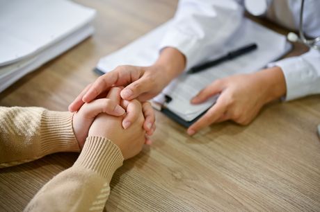 Ein Arzt in einem weißen Kittel spricht empathisch mit einem Patienten, indem er dessen Hand hält, mit Fokus auf die ineinandergreifenden Hände auf einem Holztisch.