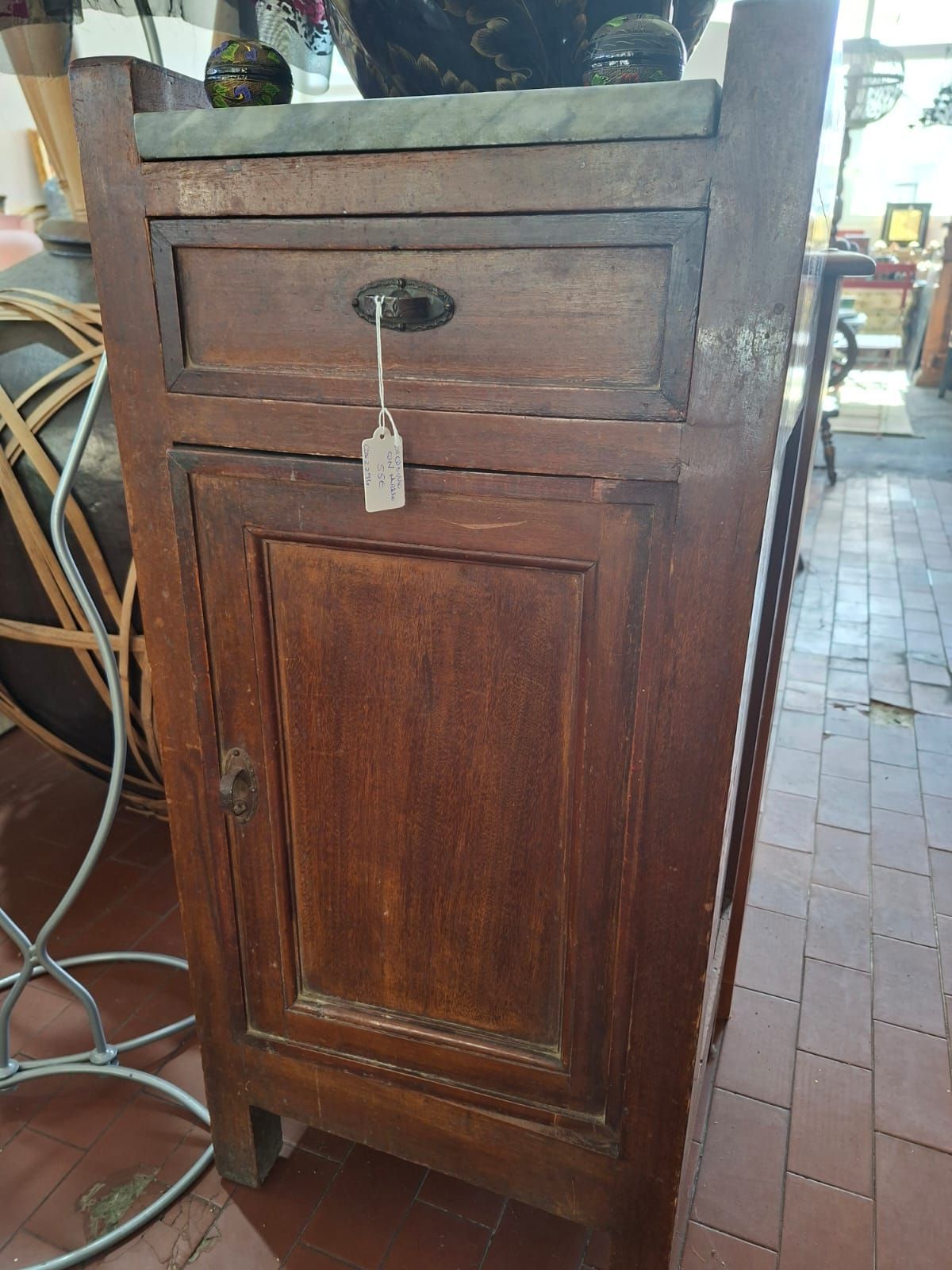 Un comodino vintage in legno con un singolo cassetto con targhetta e un'anta inferiore, sormontato da un piano in pietra.