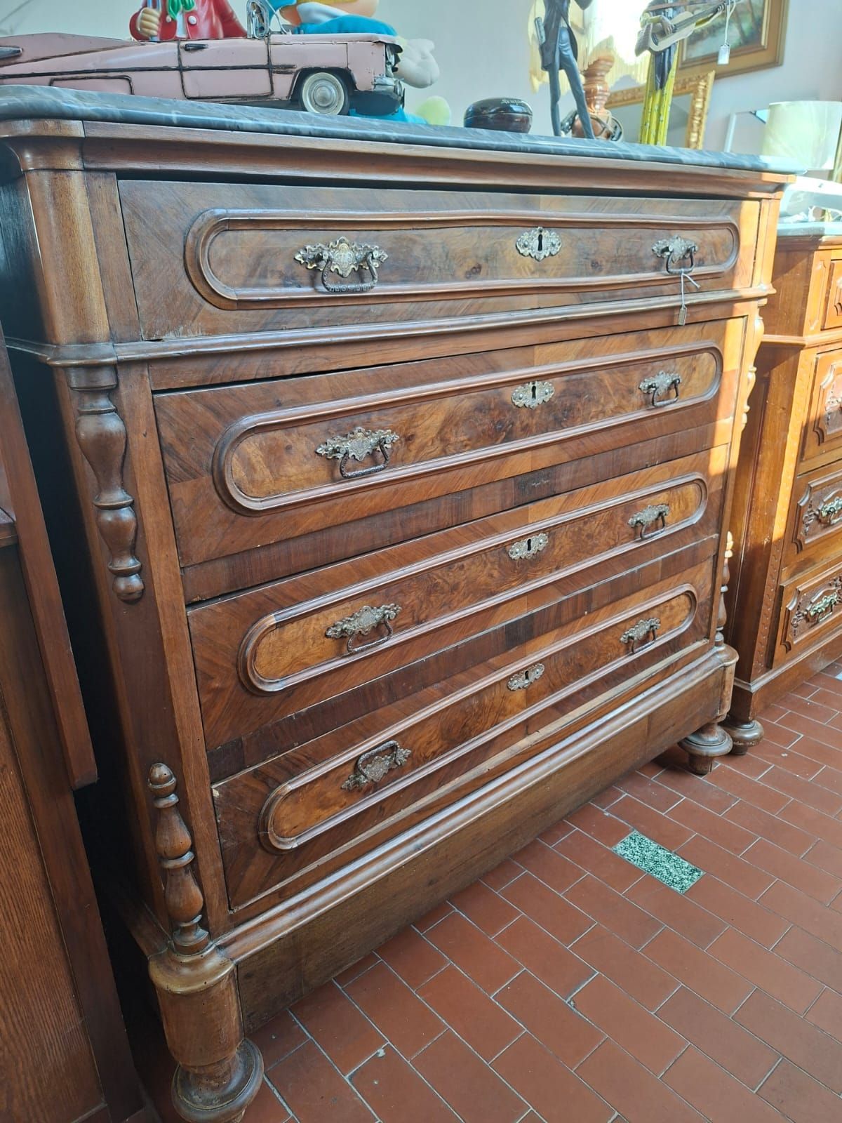 Un'alta cassettiera antica in legno scuro con quattro cassetti e piano in marmo, poggiata su un pavimento piastrellato di rosso.