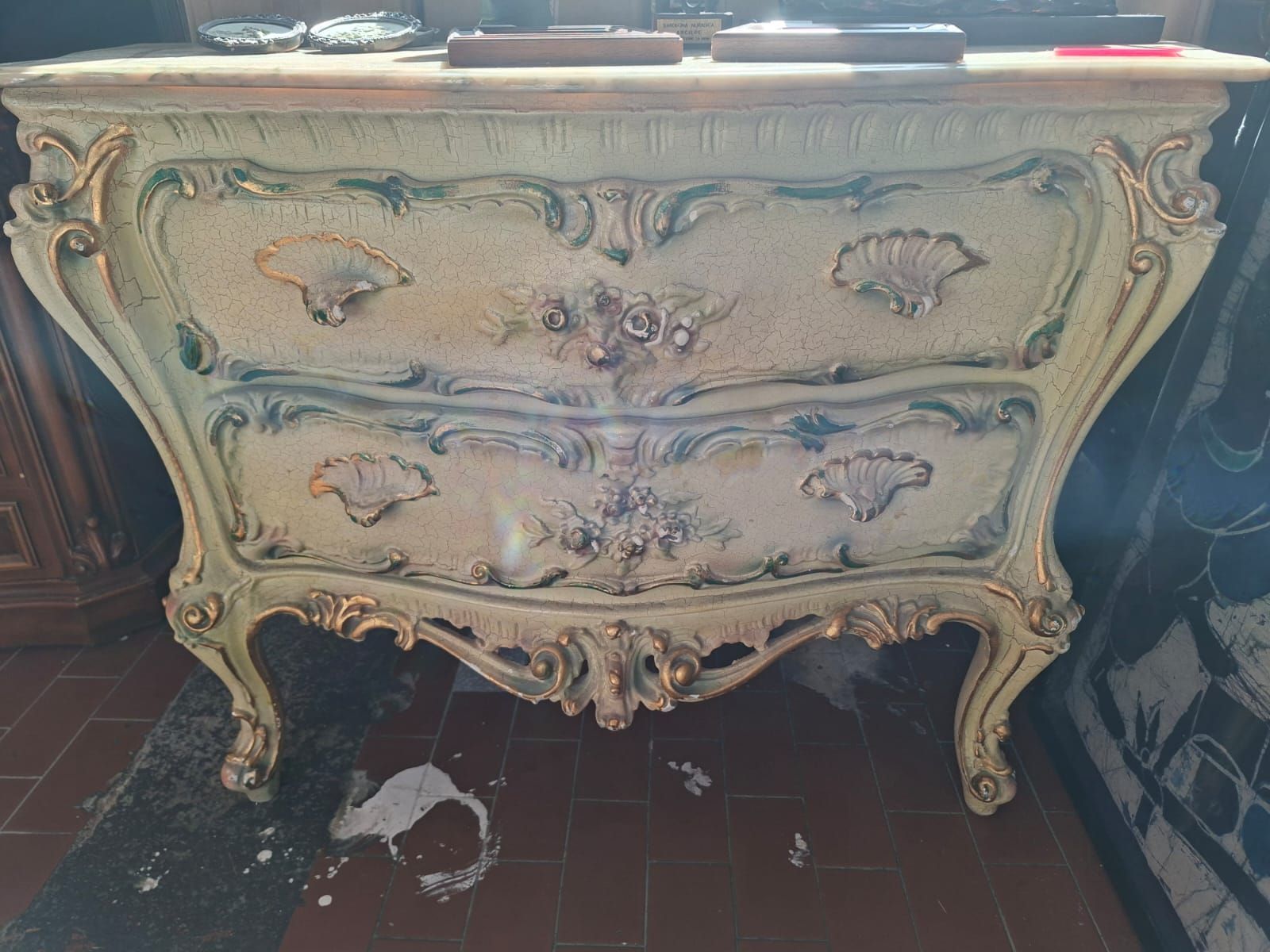 Una cassettiera elaborata, color bianco sporco, a due cassetti, con dettagli in foglia d'oro, bordi smerlati e intagli floreali su gambe curve.