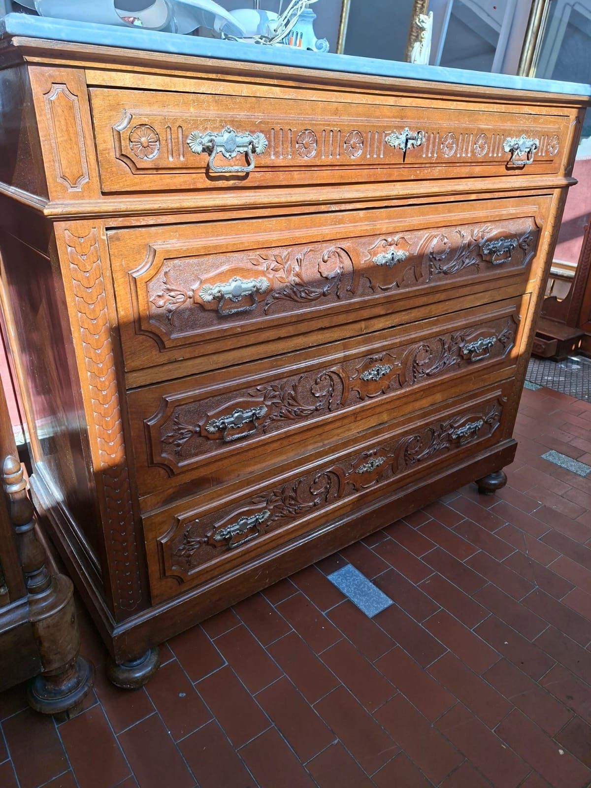 Un antico comò in legno con intagli elaborati, ferramenta in metallo e piano in pietra, poggiato su un pavimento piastrellato.