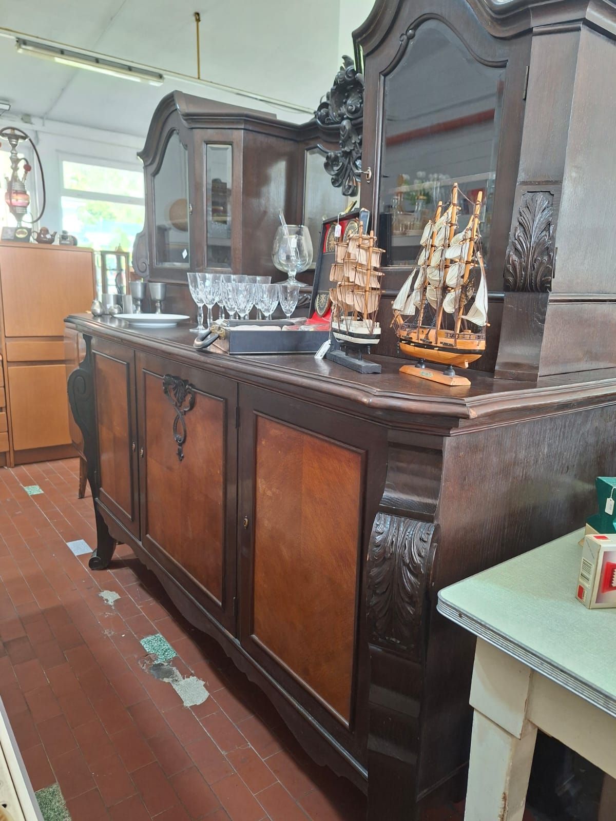 In una sala espositiva di un negozio si trova una credenza in legno scuro riccamente decorata, con una vetrina sovrastante su cui sono esposti modellini di navi e oggetti in vetro.