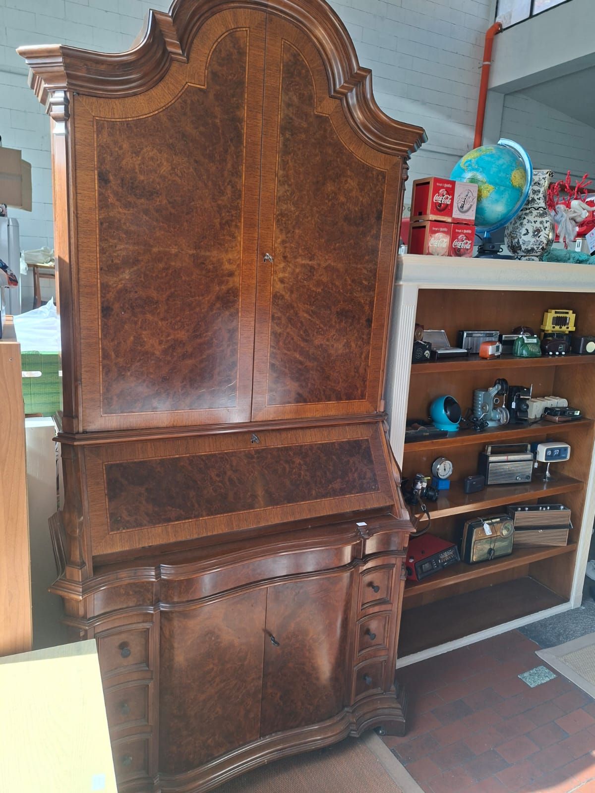 Una scrivania da segretaria alta e decorata in legno, con piano curvo e impiallacciatura in radica di noce, posta accanto a una mensola con piccoli oggetti.