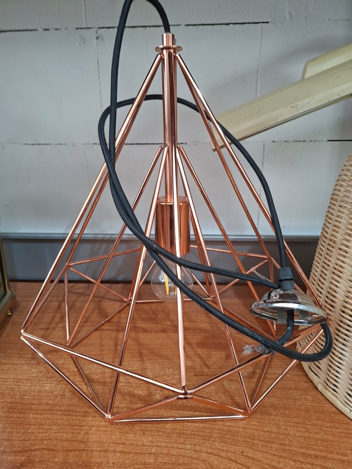 Una lampada a sospensione geometrica color rame con cavo nero poggia su una superficie di legno.