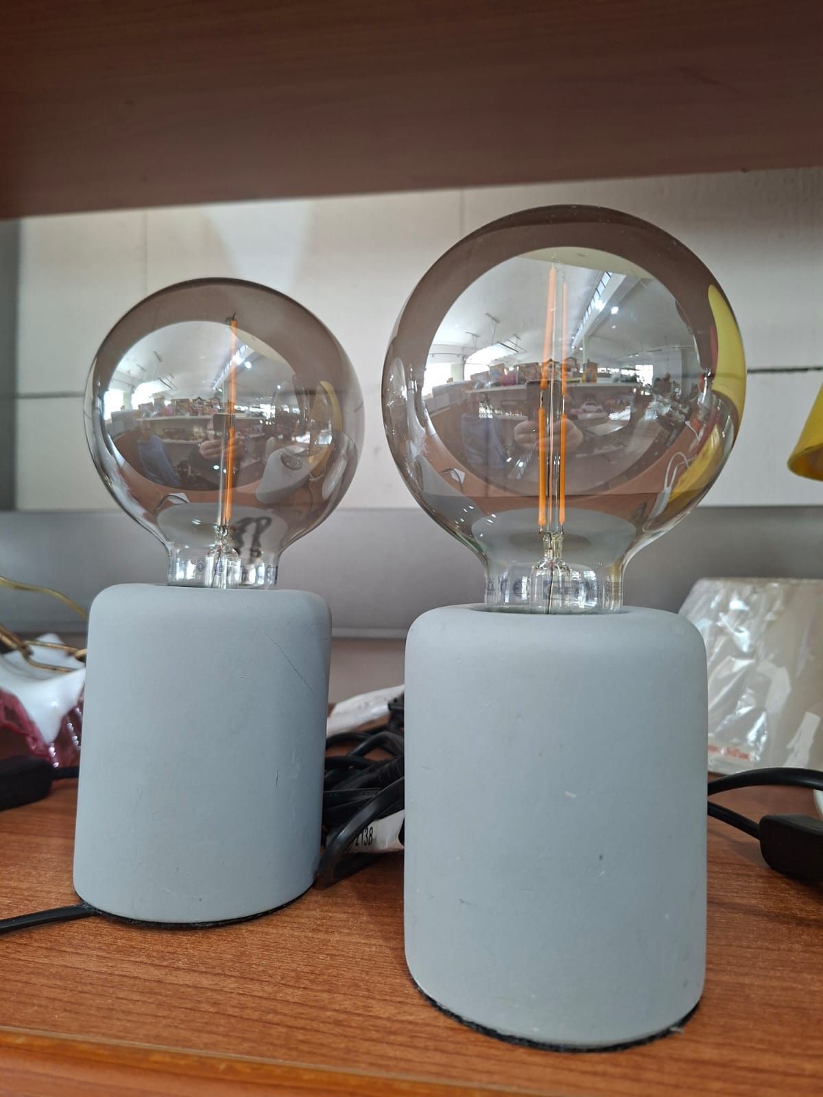 Due lampade da tavolo minimaliste con basi in cemento grigio e grandi lampadine rotonde in stile Edison vintage, posizionate su una mensola di legno.