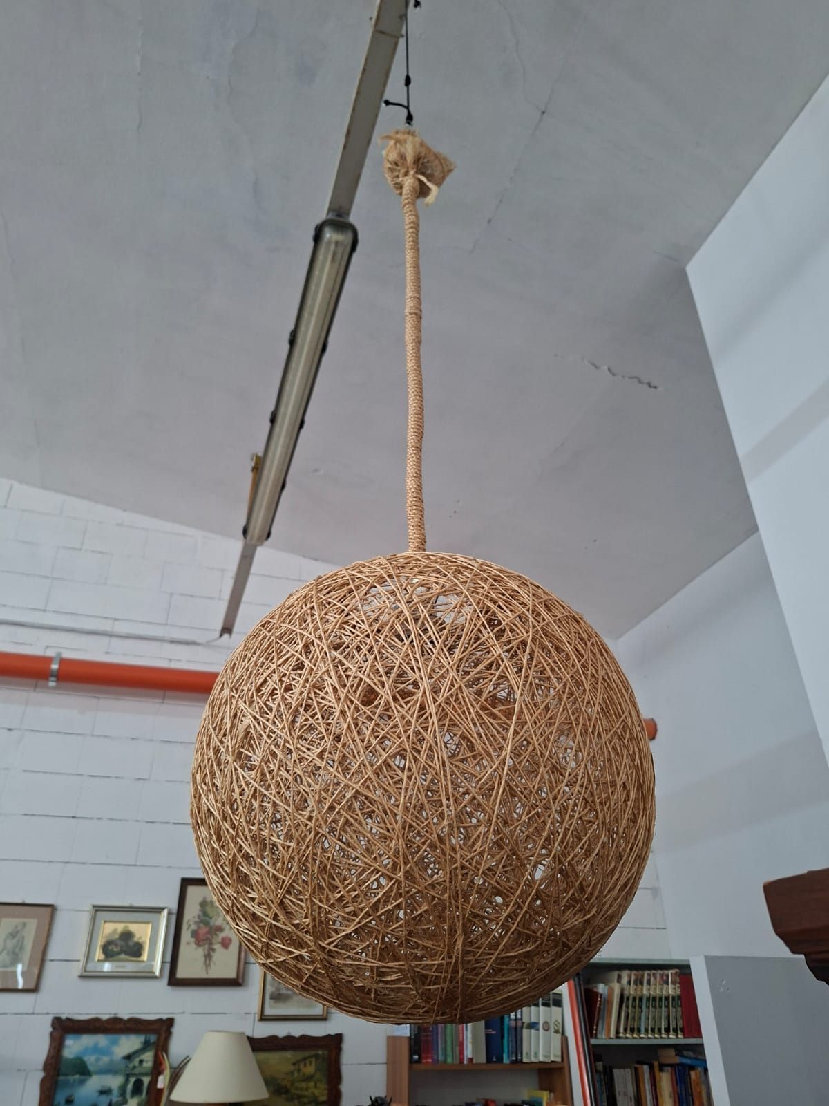 Una lampada a sospensione sferica realizzata in spago intrecciato di colore naturale, sospesa tramite un cavo abbinato a una parete bianca.