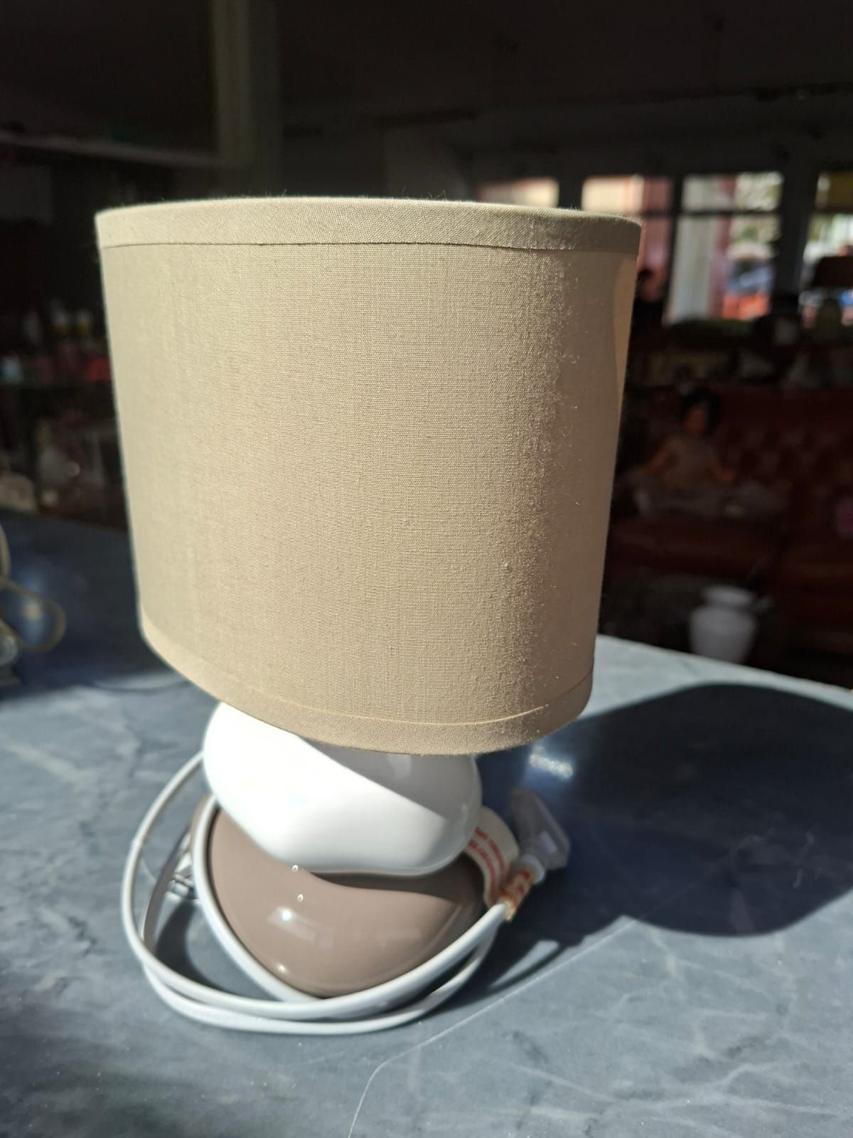 Una piccola lampada da tavolo moderna con paralume in tessuto color cuoio, poggiata su una base in ceramica bicolore a forma di ciottolo, su una superficie grigia.