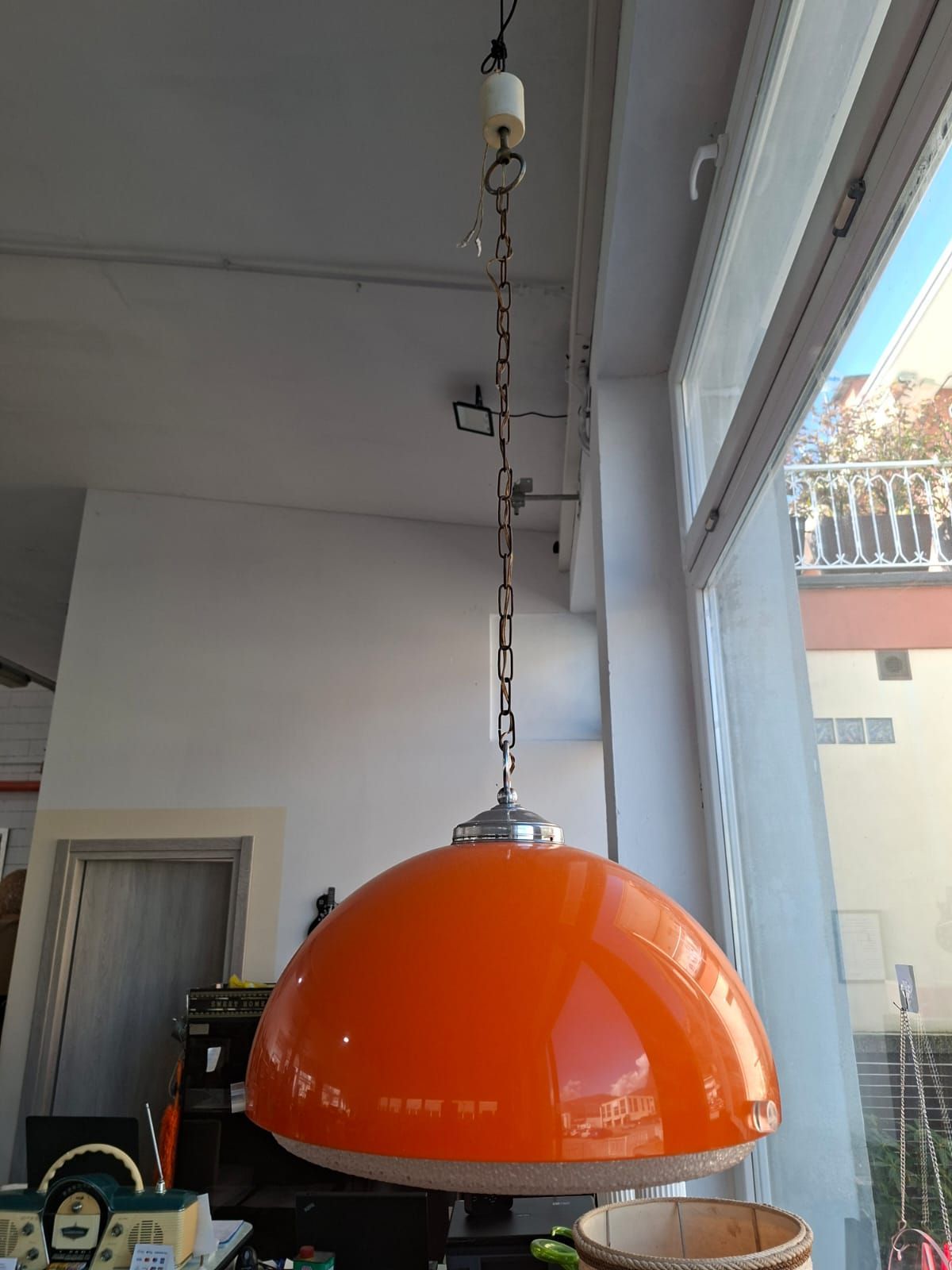 In un ambiente interno ben illuminato, una lampada a sospensione a cupola di colore arancione brillante, in stile moderno di metà secolo, pende da una catena d'argento.