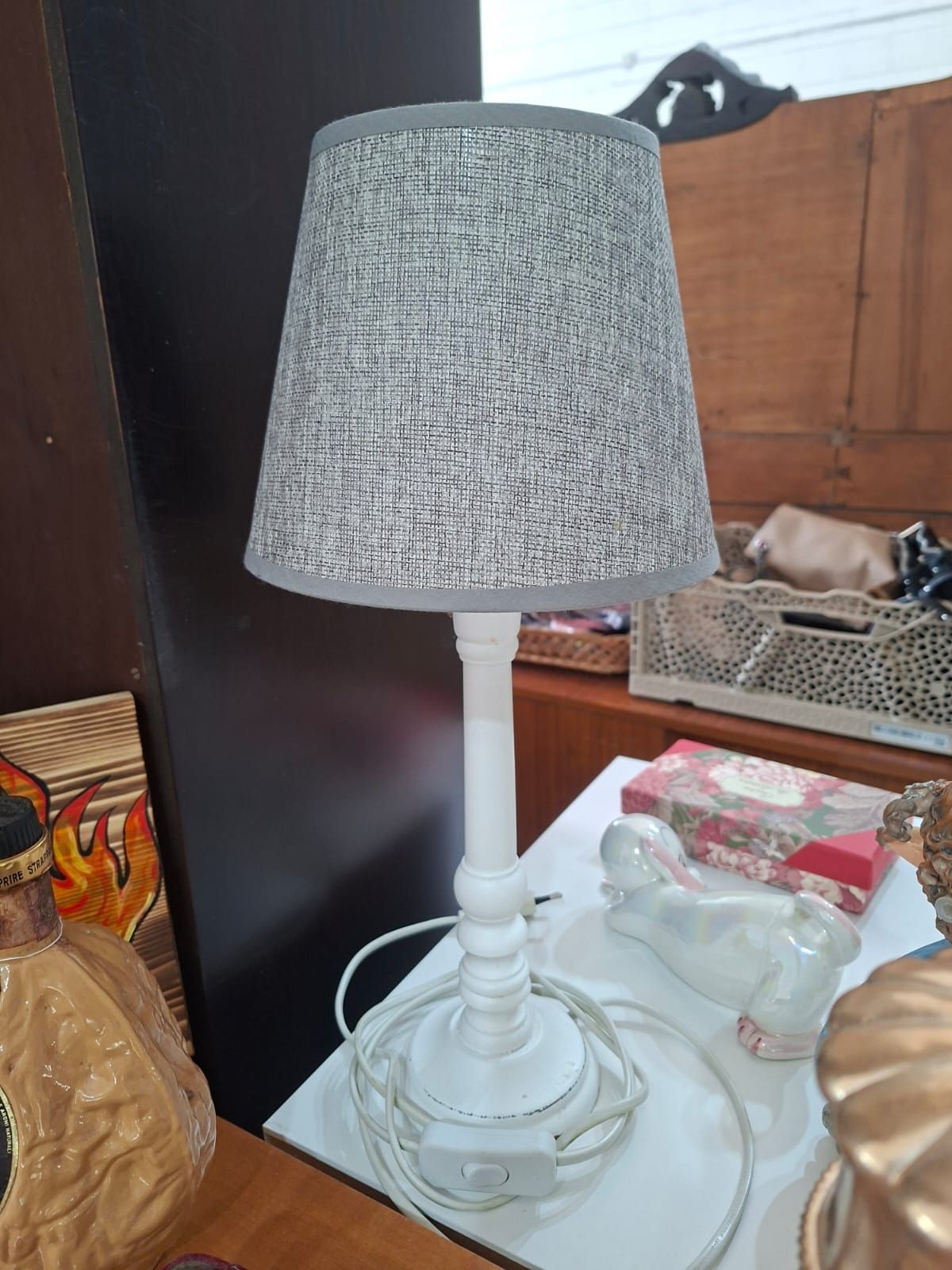 Una lampada da tavolo bianca con paralume in tessuto grigio testurizzato, appoggiata su un tavolo tra altri oggetti.