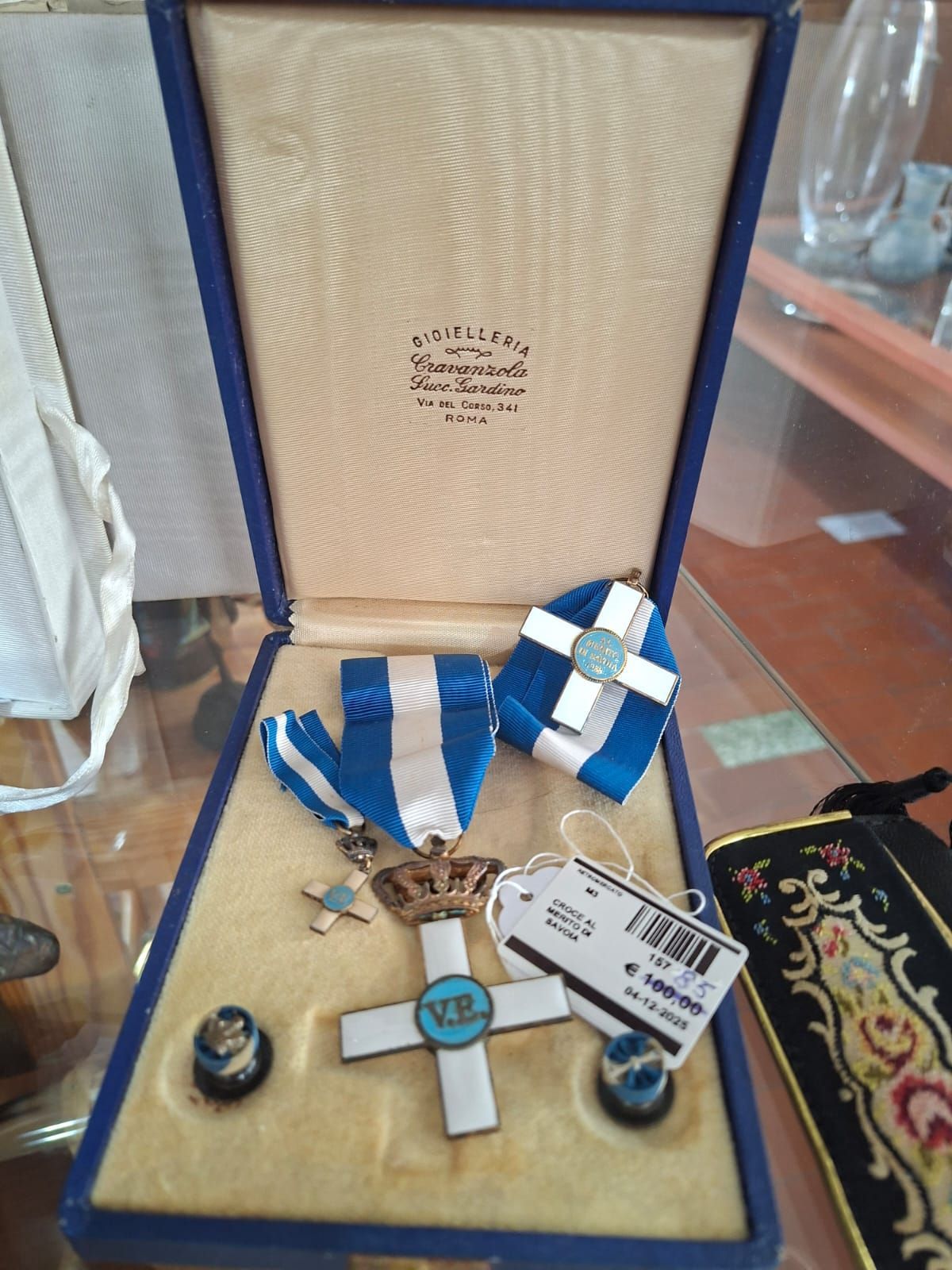 Un cofanetto blu aperto contenente una decorazione reale greca, comprendente un ciondolo a croce, un nastro e due bottoni.