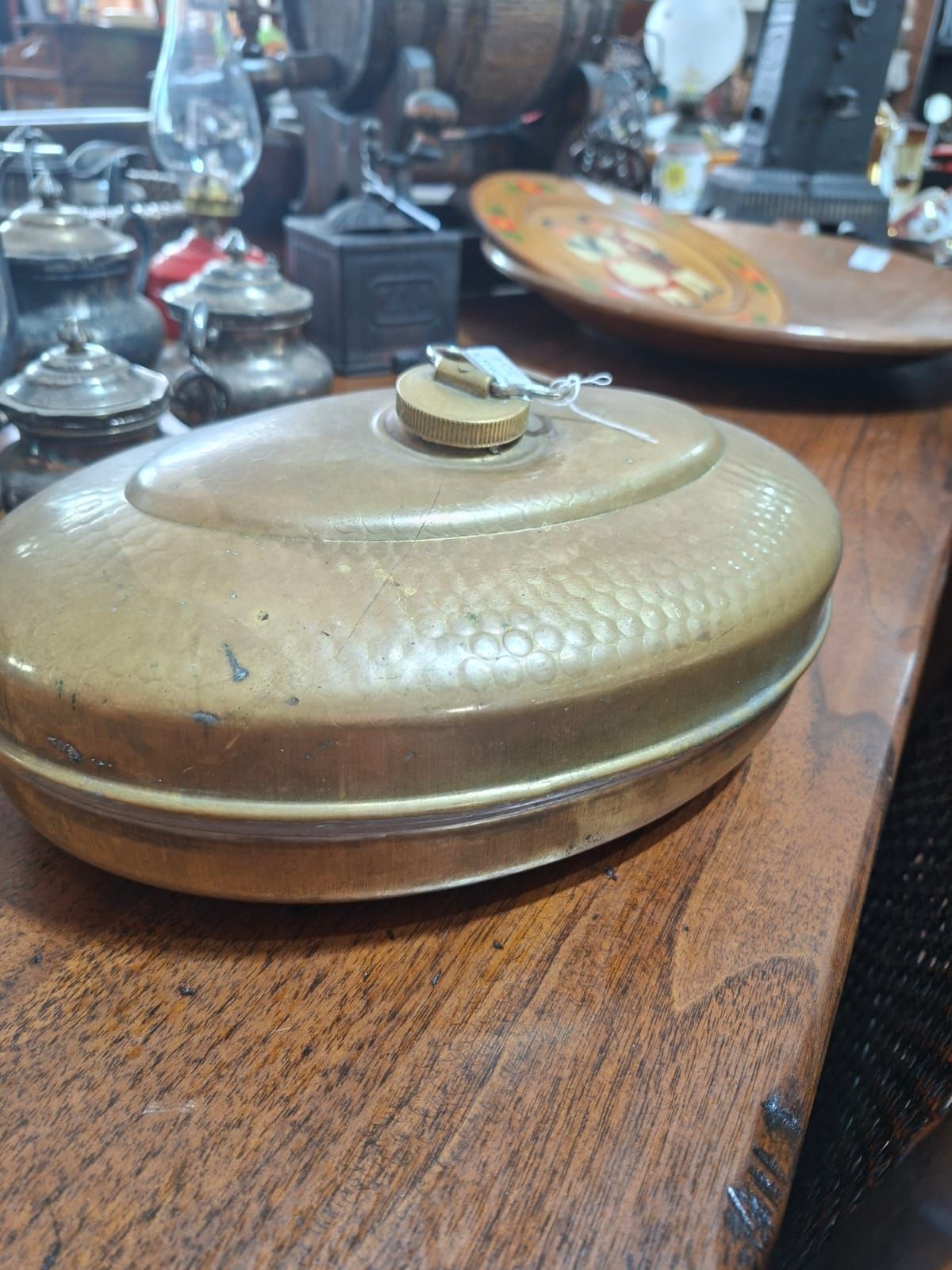 Una borsa dell'acqua calda antica, ovale, in metallo martellato, con tappo a vite, appoggiata su una superficie di legno in un negozio di antiquariato.