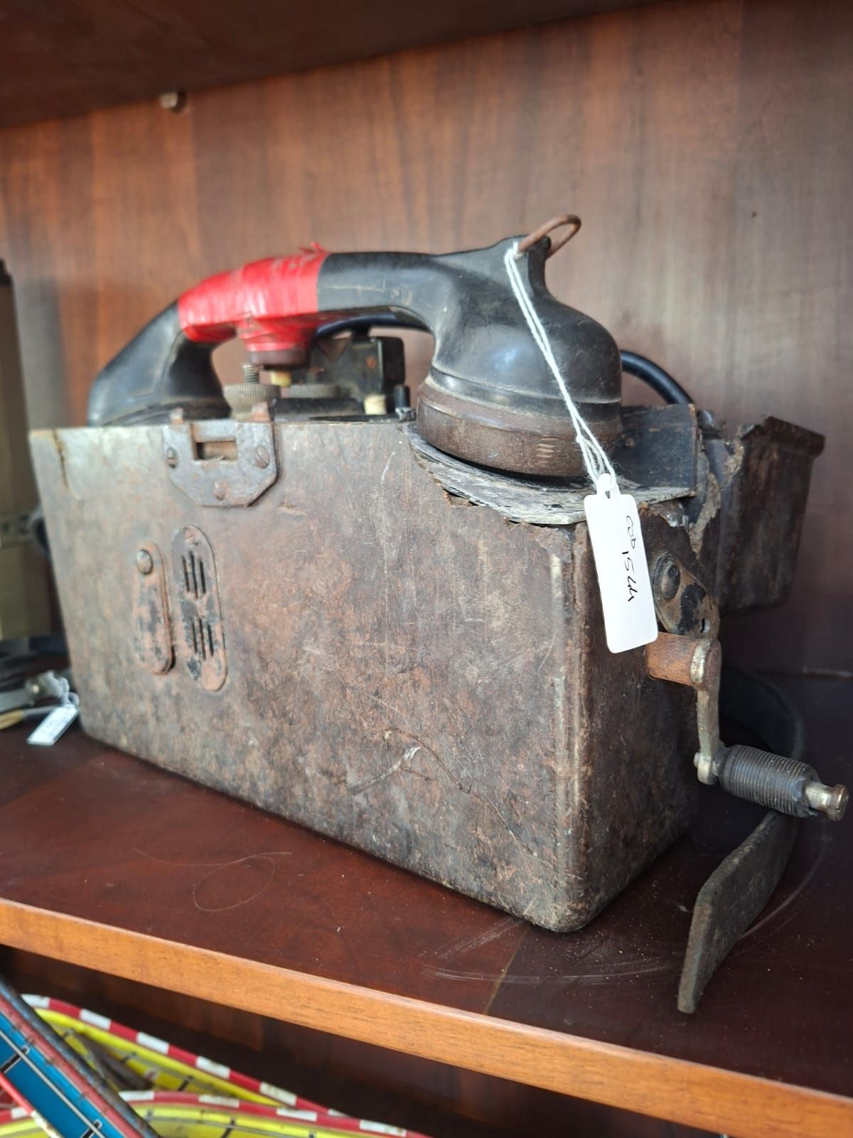 Un vecchio telefono da campo in metallo, consumato dal tempo, con manovella e ricevitore appoggiato su uno scaffale di legno.