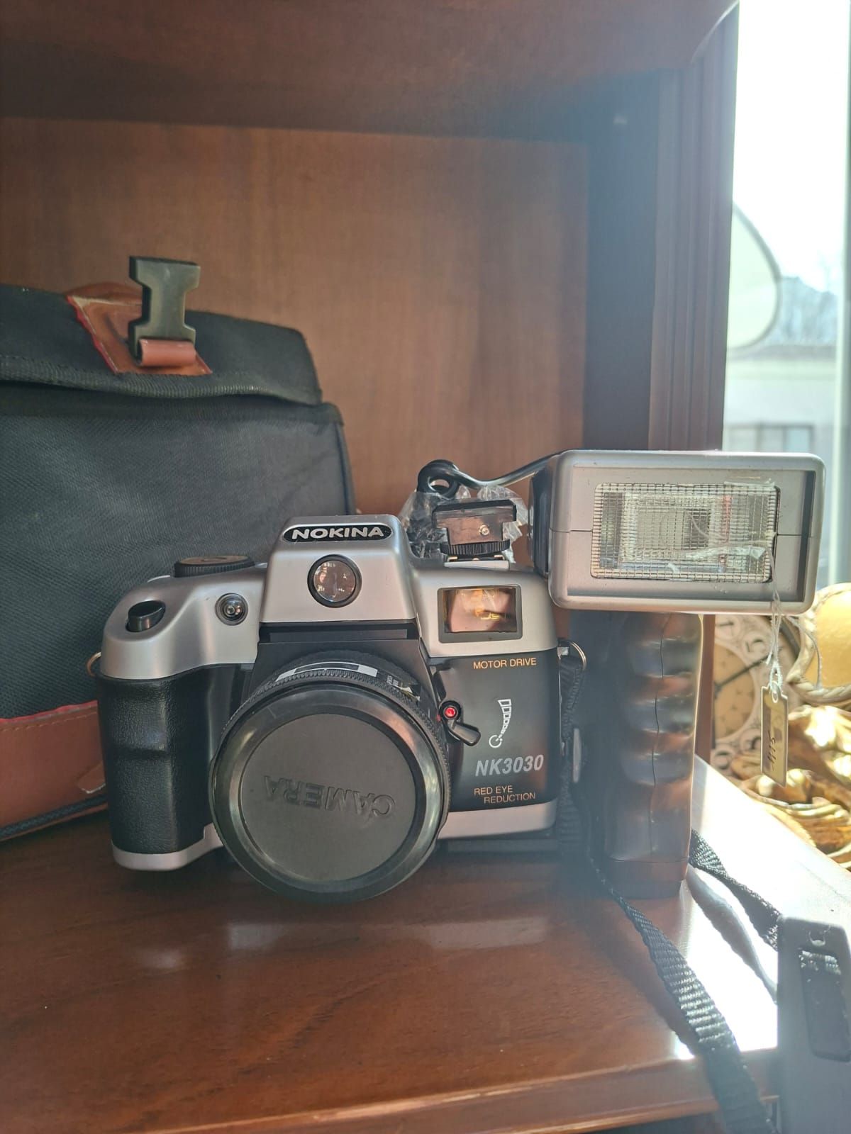 Una fotocamera zoom Olympus AZ-300 color argento con flash incorporato, appoggiata su uno scaffale di legno accanto a una borsa fotografica nera.