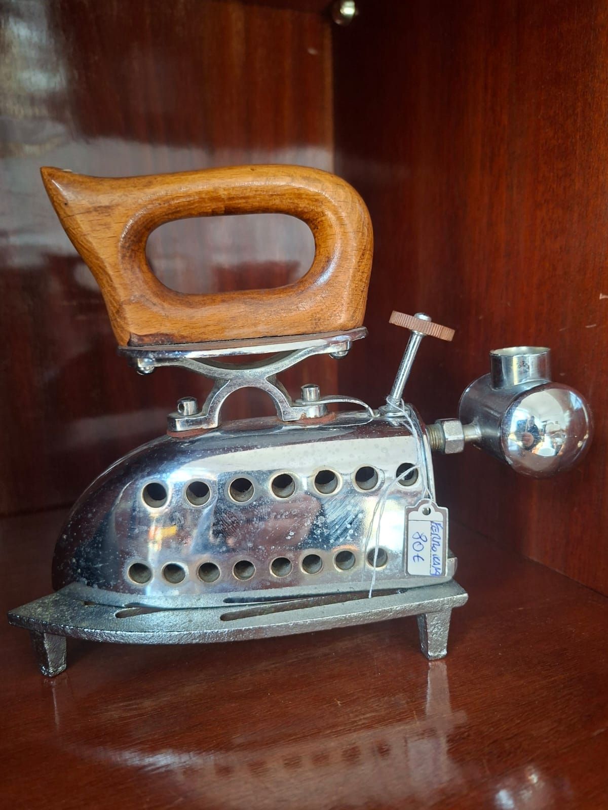 Un ferro da stiro a carbone vintage in cromo con manico in legno e piccolo camino, appoggiato su un supporto metallico all'interno di un armadietto.