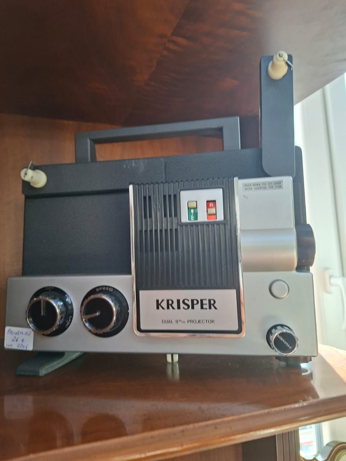 Un proiettore cinematografico vintage Krisper grigio e nero, completo di manopole e interruttori di controllo, appoggiato su una mensola di legno.