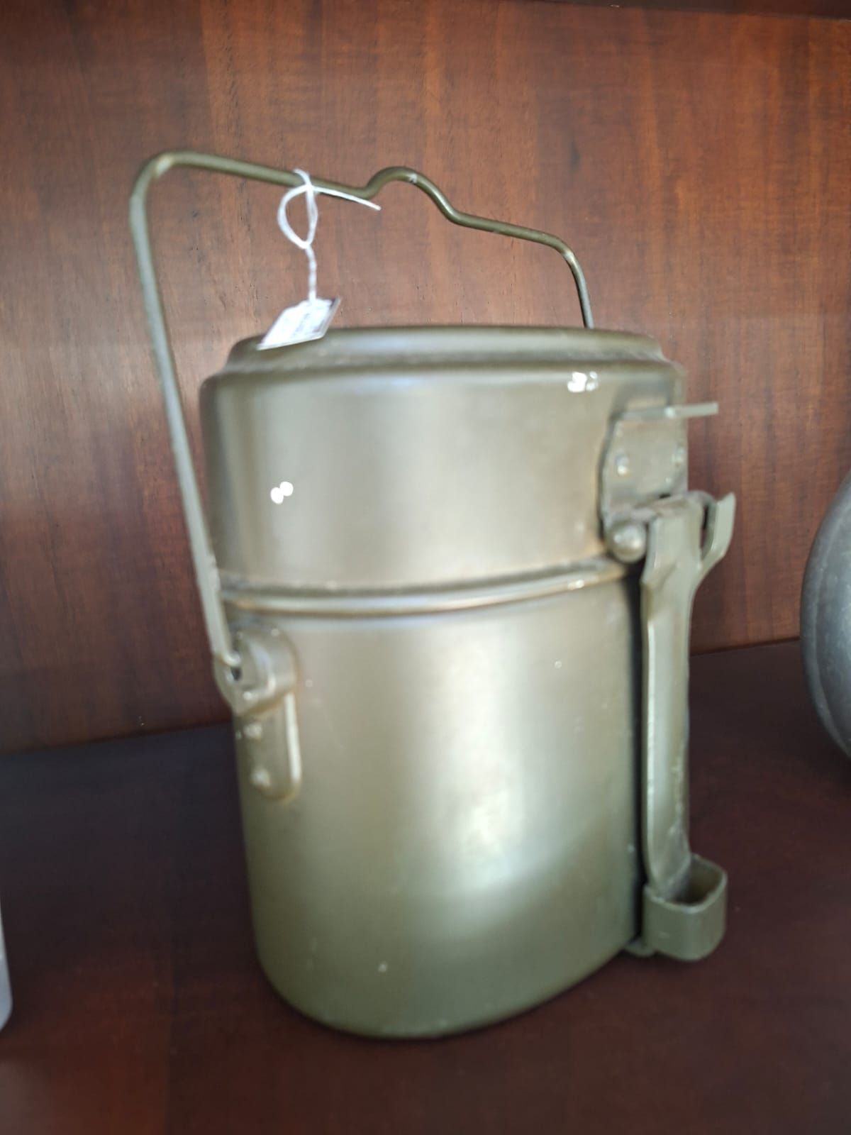 Una gavetta militare in metallo color verde oliva con manico in filo metallico, appoggiata su una superficie di legno scuro.