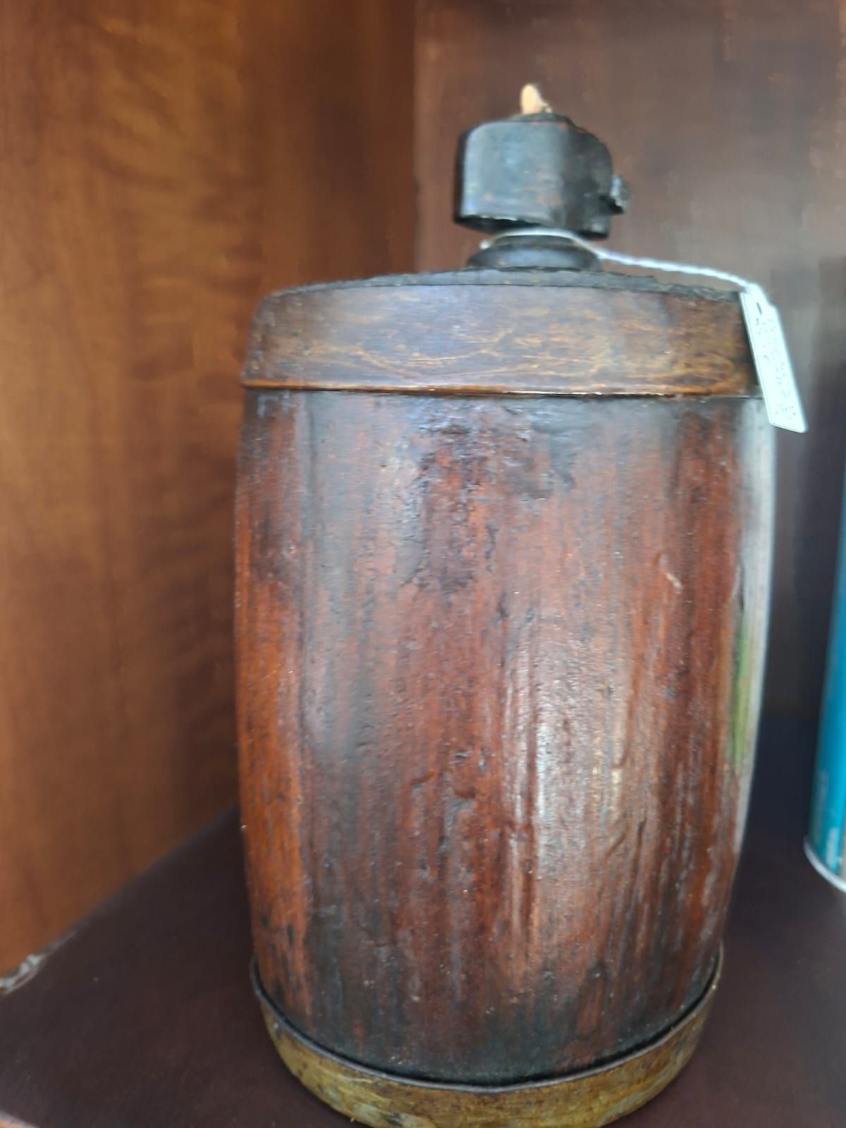 Un contenitore cilindrico in legno con finitura scura, coperchio nero e base in metallo, appoggiato su uno scaffale.