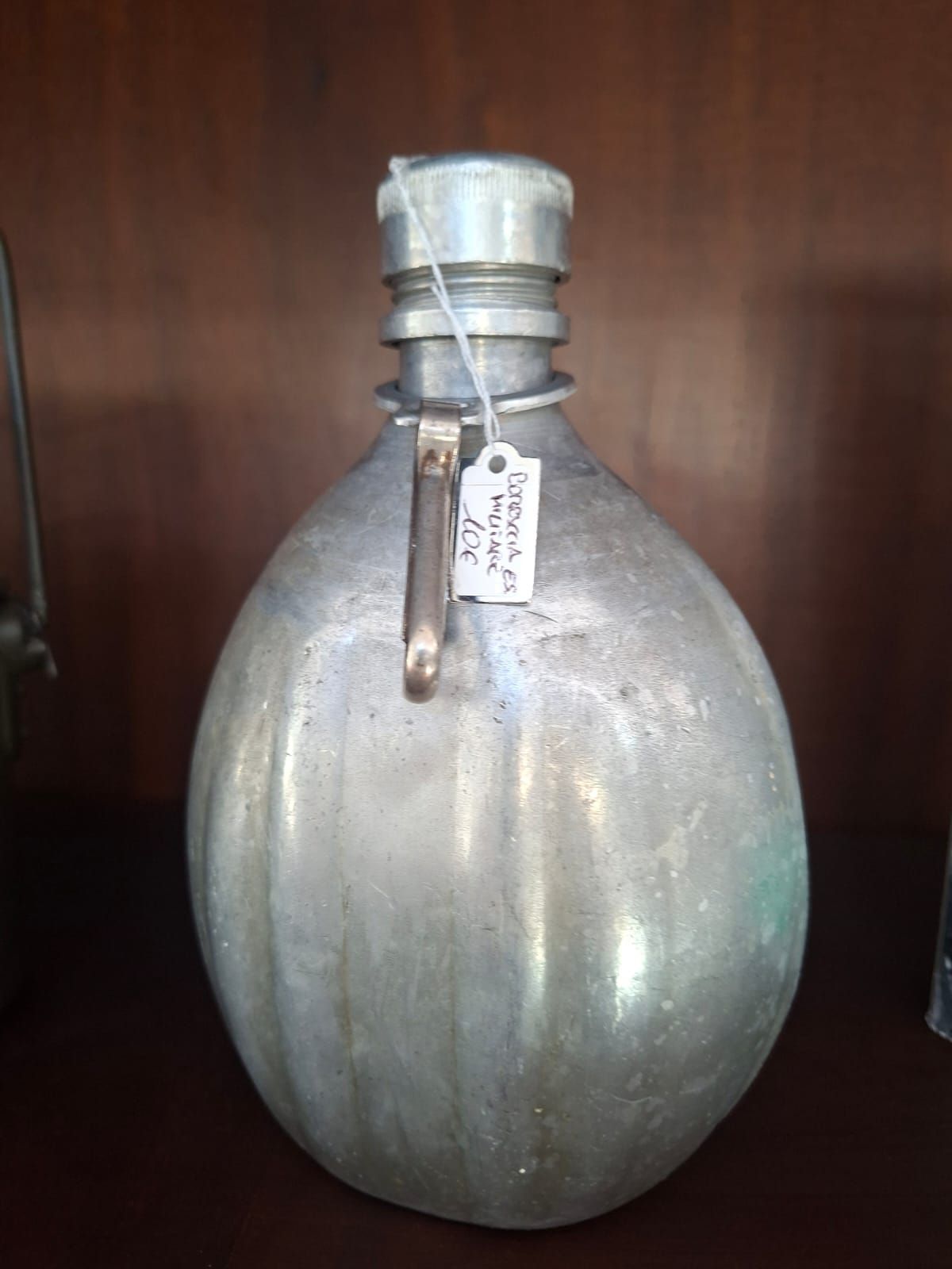 Una borraccia vintage in alluminio a coste con tappo in metallo e una piccola etichetta bianca con il prezzo, su uno sfondo di legno scuro.
