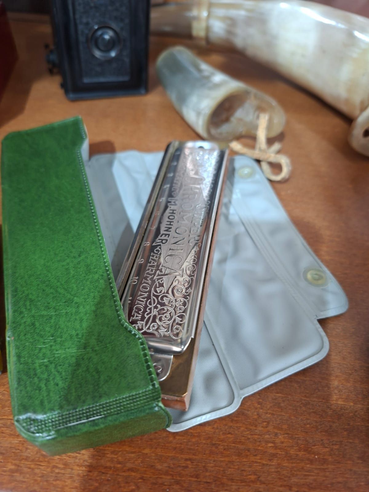 Un'armonica d'argento riccamente decorata si trova accanto a una custodia di velluto verde, un panno grigio e uno strumento musicale su un tavolo di legno.