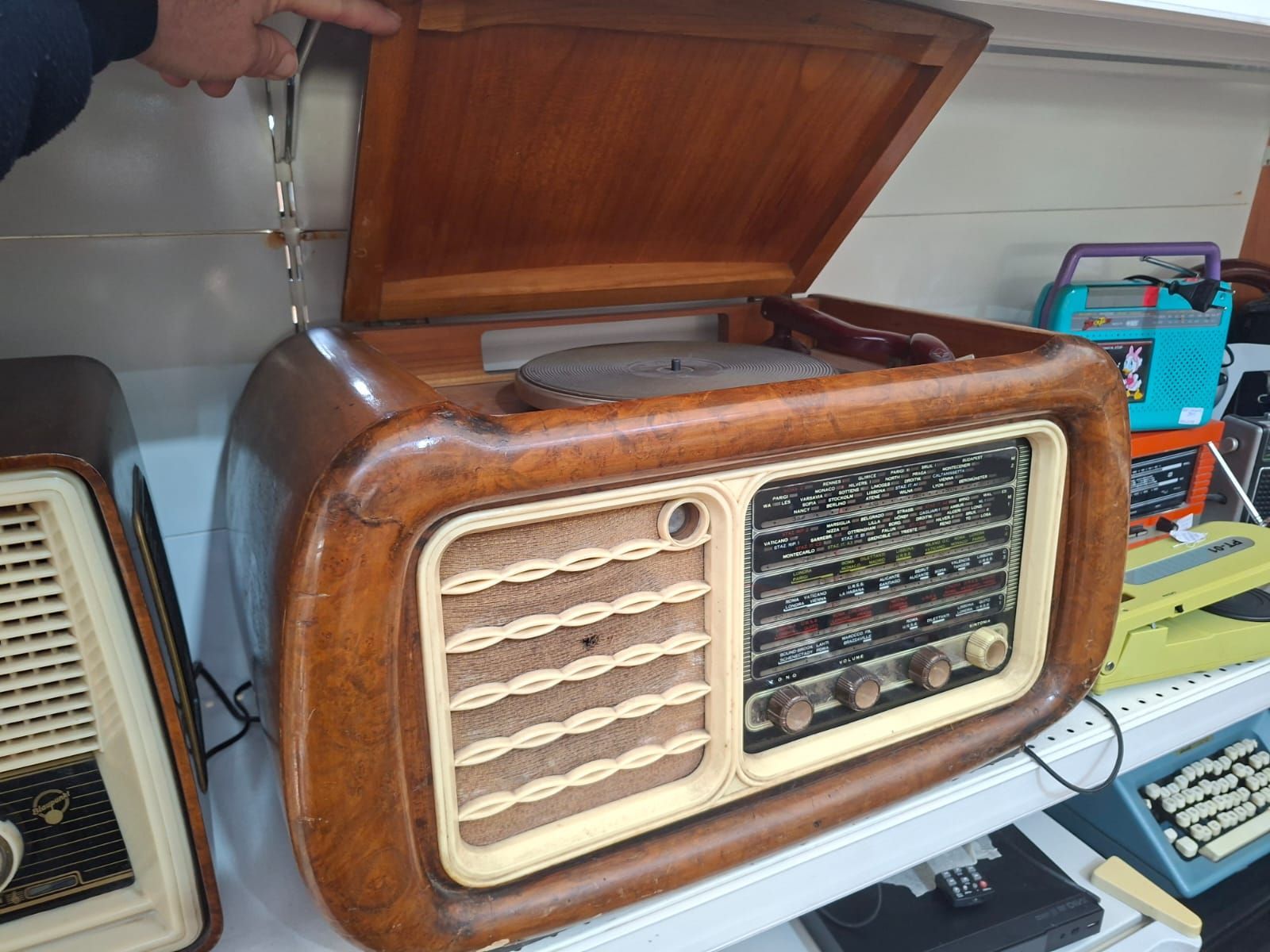 Un vecchio radiofonografo in legno con coperchio ribaltabile, posizionato su uno scaffale di un negozio accanto ad altri apparecchi elettronici d'epoca.