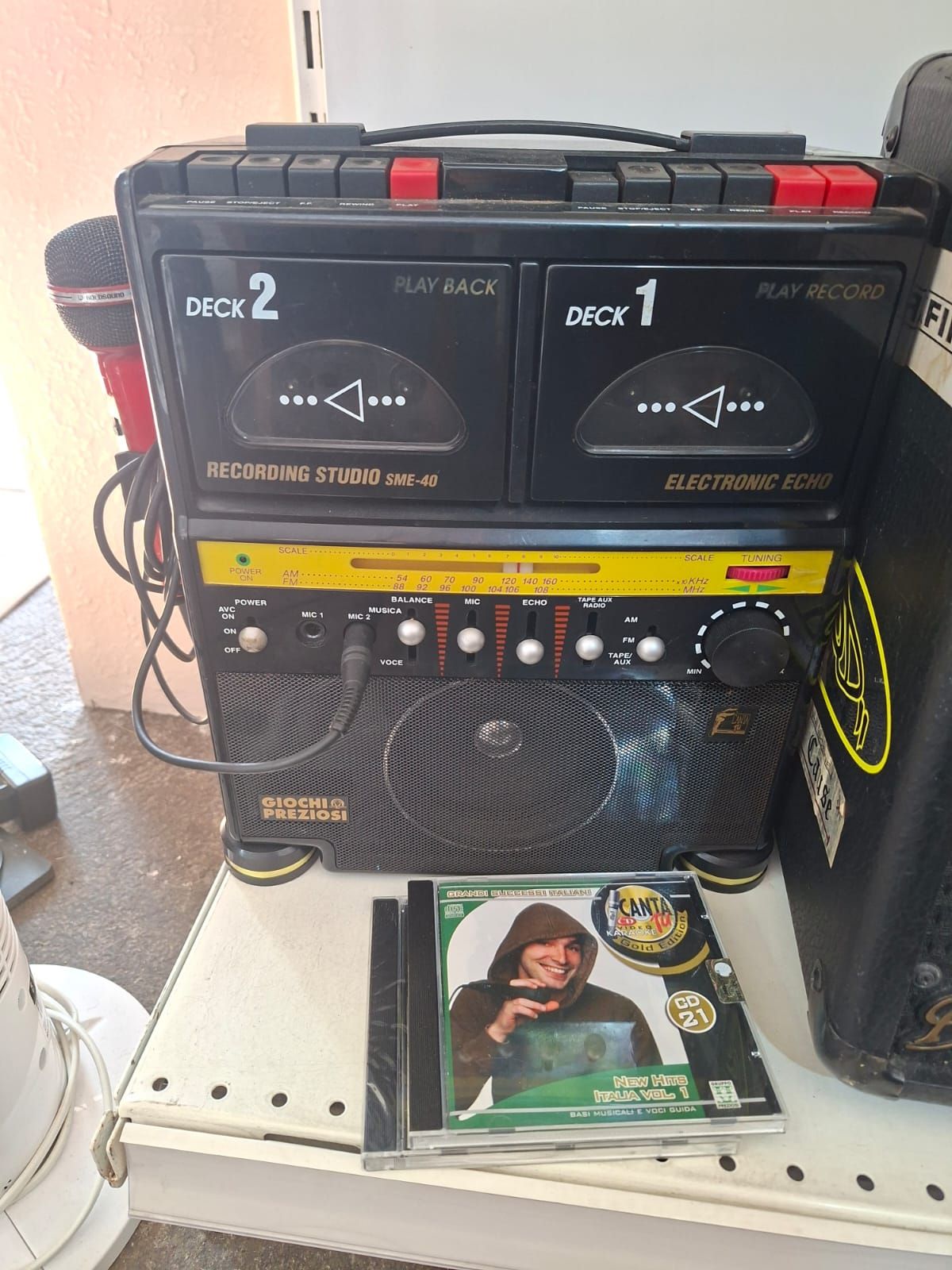 Su uno scaffale di un negozio è appoggiato un registratore a cassette portatile a doppio deck di colore nero, con una custodia per CD musicali posizionata davanti ad esso.