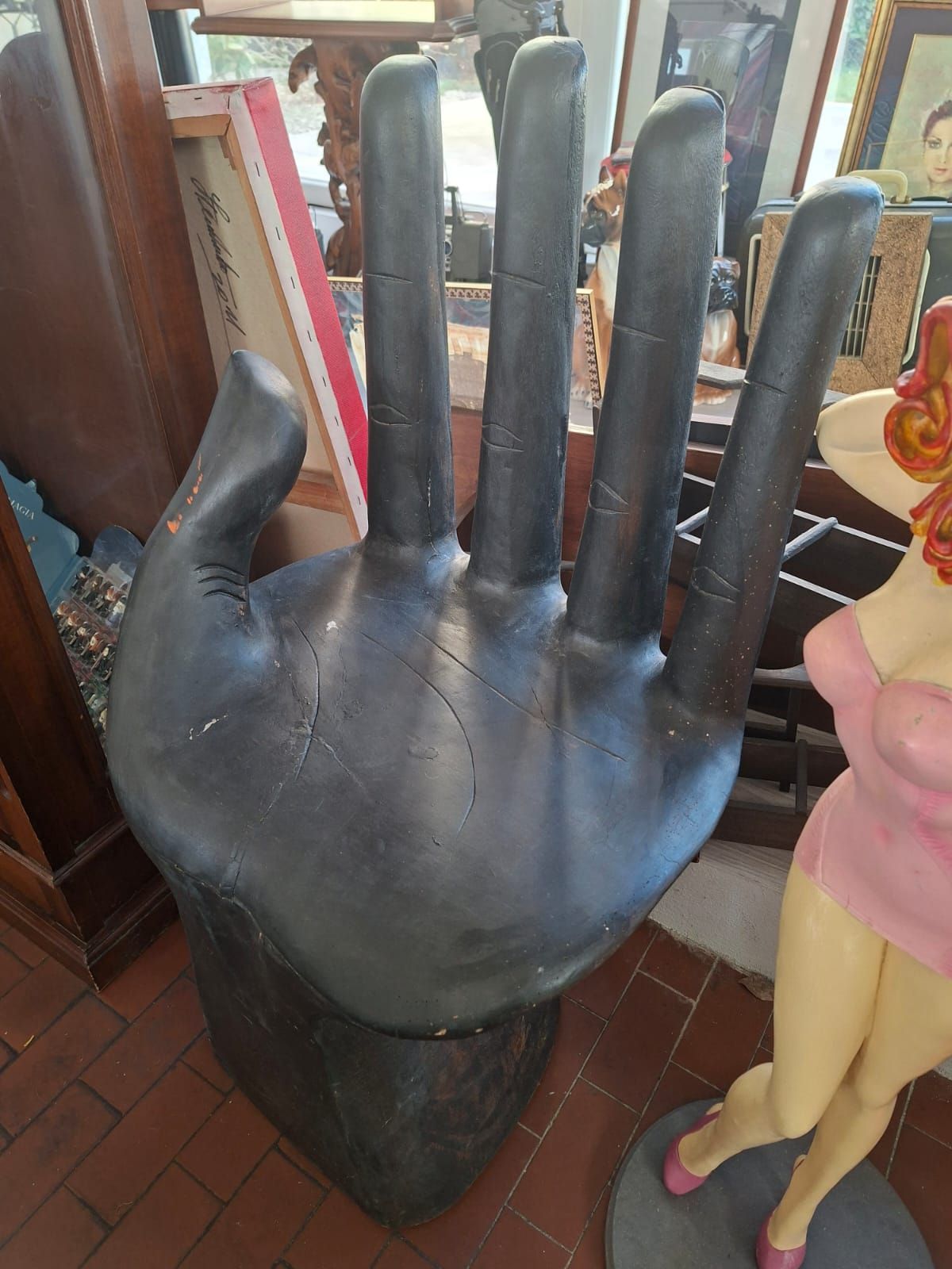 In uno spazio interno, accanto a un manichino, si trova una grande sedia a forma di mano di colore grigio scuro.