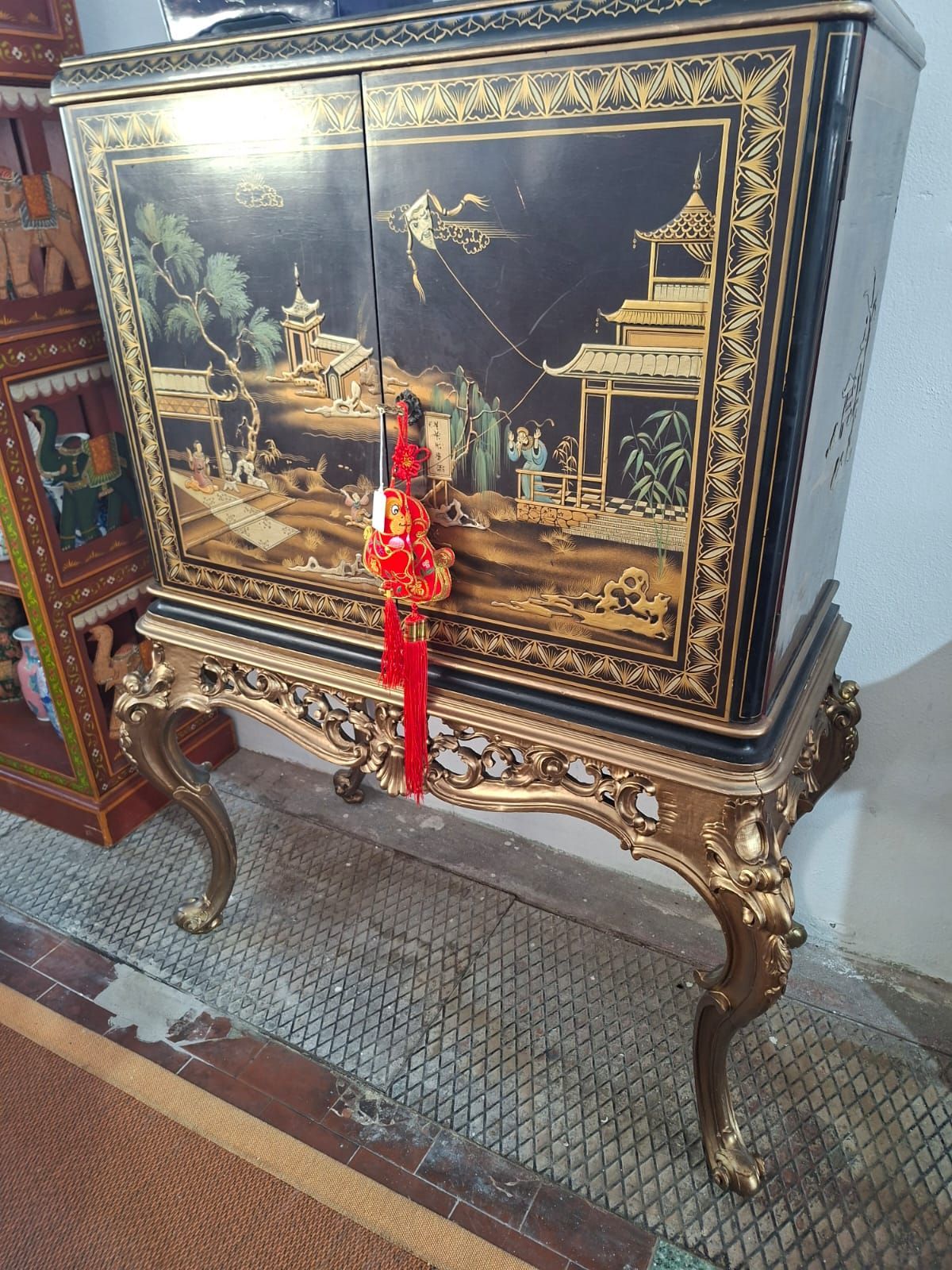 Un elaborato mobiletto laccato nero con decorazioni in stile chinoiserie dorata su una base decorativa dorata, ornato da una nappa rossa.