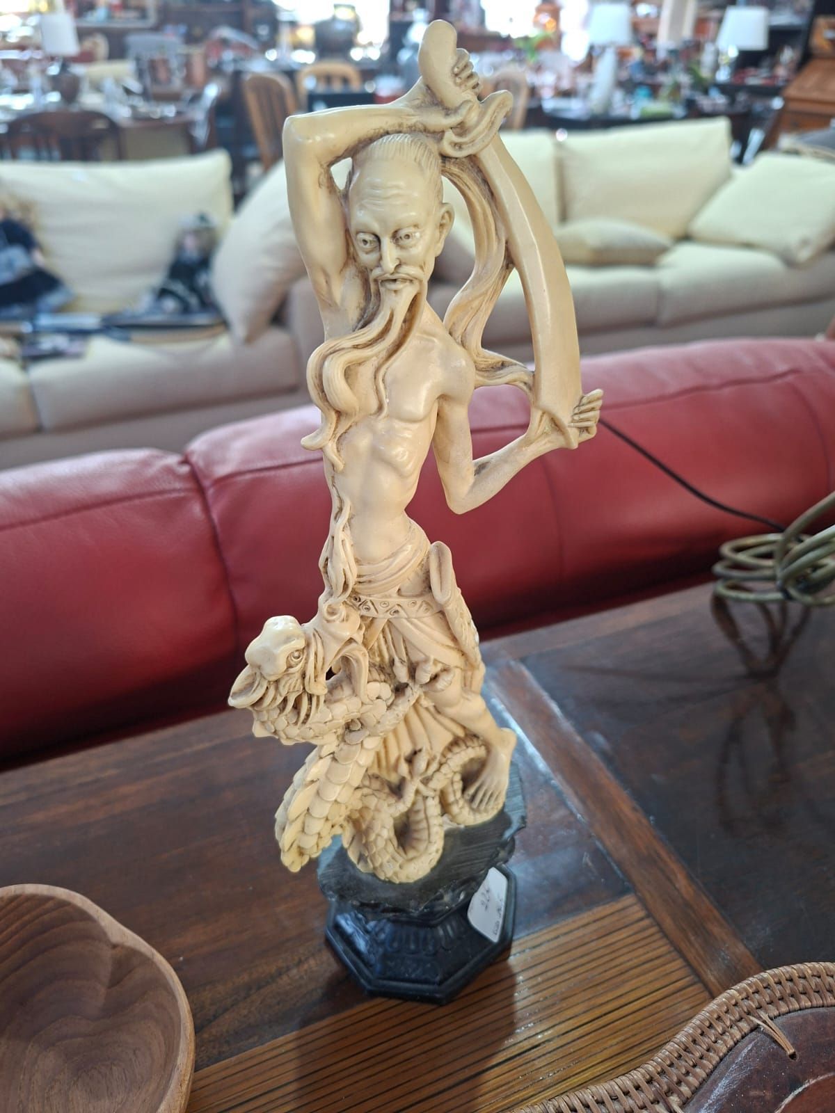 Una scultura intagliata color avorio raffigurante una figura barbuta che regge un lungo stendardo fluente, montata su una base esagonale scura.