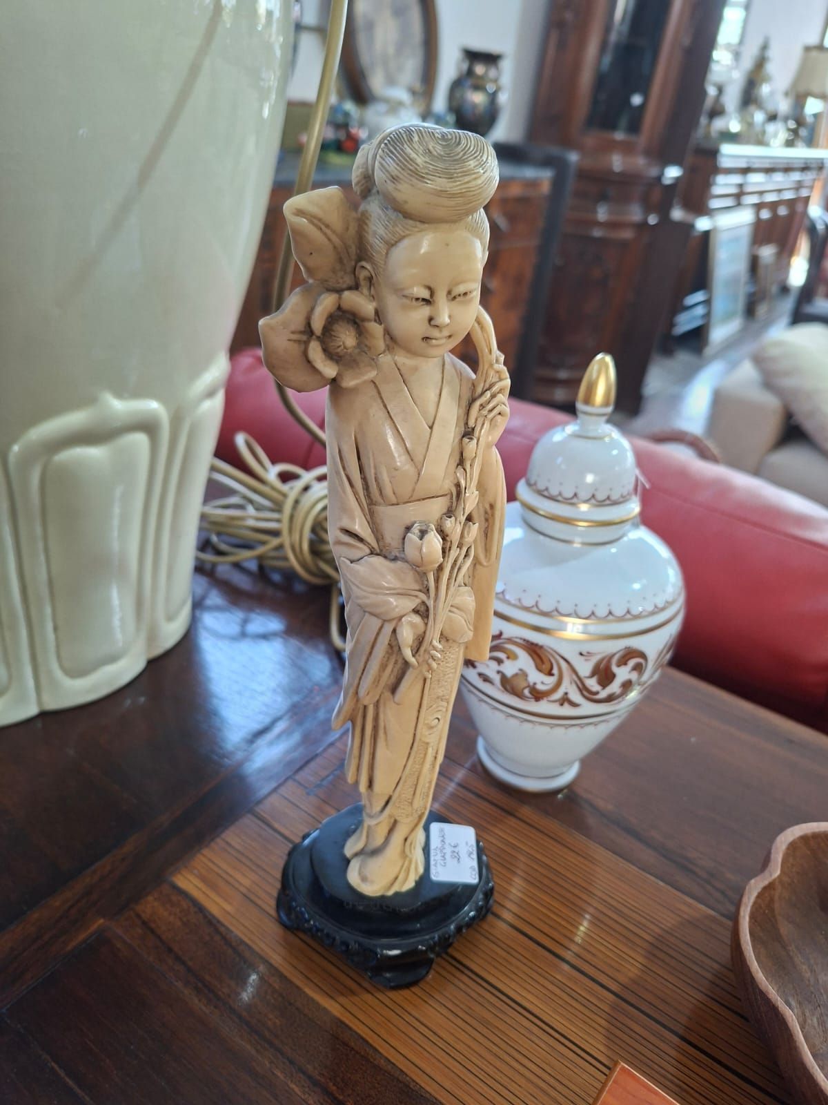 Una statuetta intagliata color bianco sporco raffigurante una figura in piedi che tiene in mano un fiore di loto, posta su una base circolare scura su un tavolo di legno.