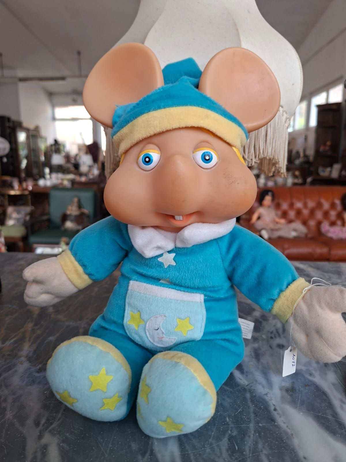 Un topolino giocattolo dal corpo morbido, con pigiama blu, decorazioni a forma di stella gialla e testa in plastica modellata, seduto su un bancone.