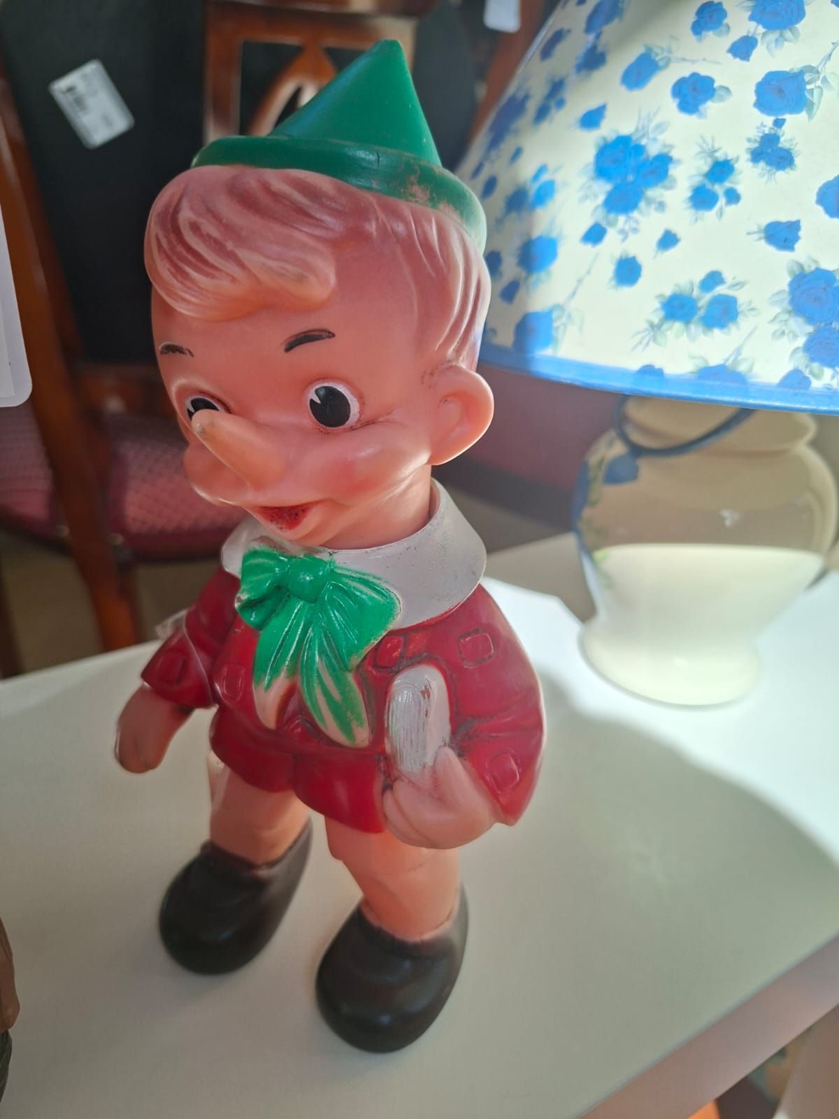 Una statuetta vintage di Pinocchio in plastica stampata, con indosso un abito rosso e un cappello verde, in piedi su una superficie bianca accanto a una lampada.