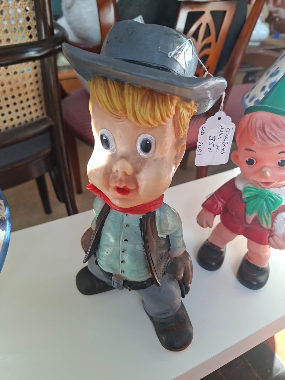 Una statuetta di un cowboy con i capelli biondi e un cappello grigio è posizionata accanto a una piccola statuetta di un elfo su un tavolo bianco.