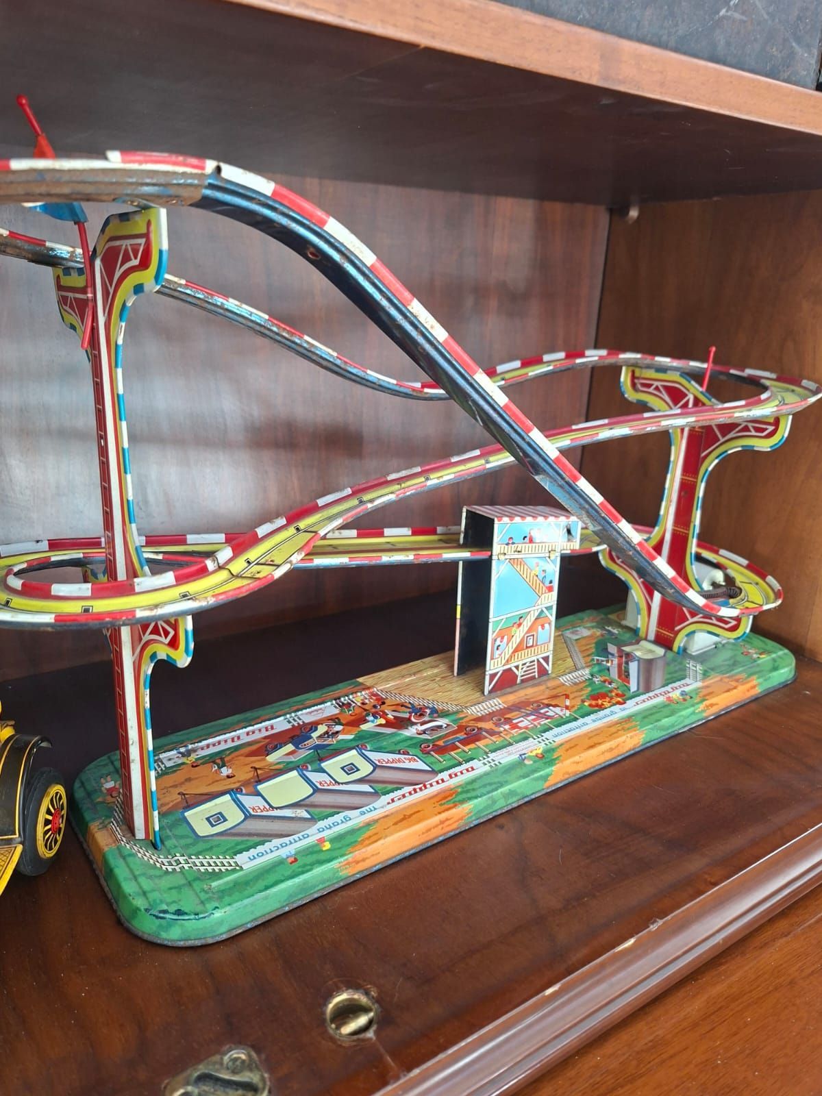 Montagne russe giocattolo vintage in latta litografata, con binari rossi e bianchi su una base verde, esposte su uno scaffale di legno.