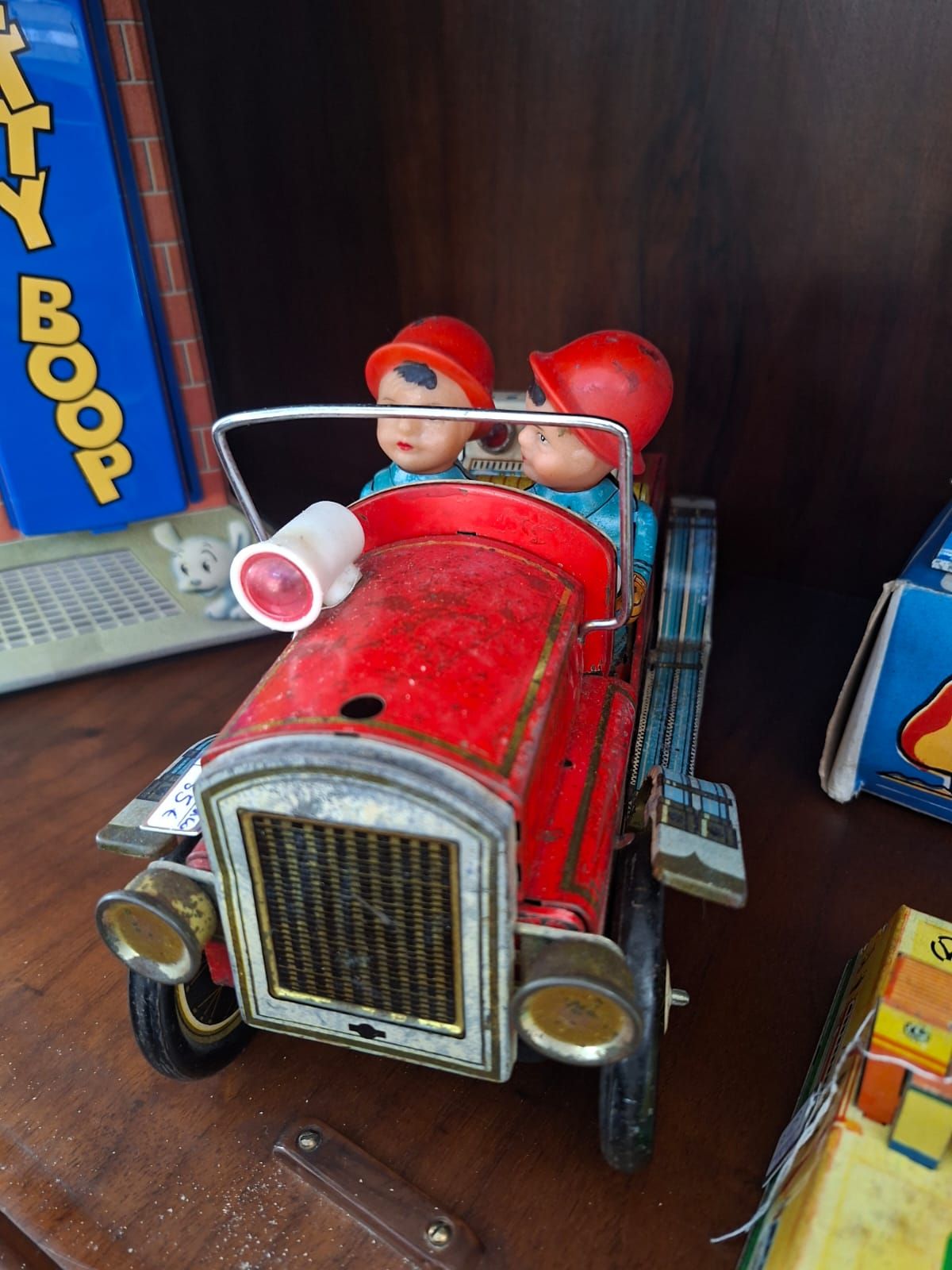 Un camion dei pompieri giocattolo d'epoca in latta con due personaggi con elmetto rosso, esposto su uno scaffale di legno accanto ad altre scatole di giocattoli.