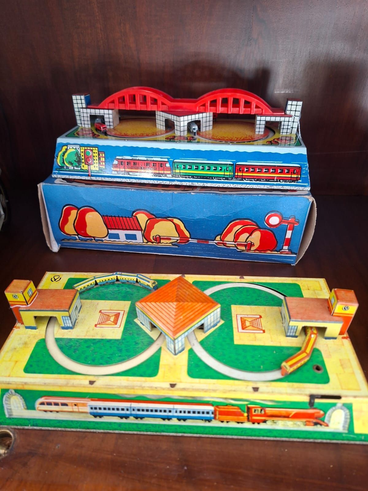 Due set di trenini giocattolo vintage in cartone con binari, gallerie e un ponte rosso stampati, esposti su una superficie di legno.