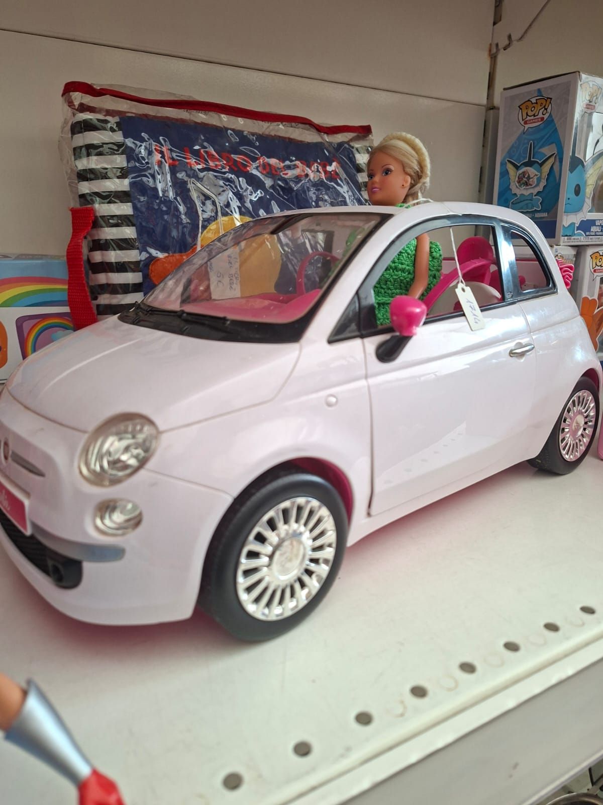Su uno scaffale di un negozio, una bambola è seduta al posto di guida di una piccola macchinina Fiat rosa pallido.