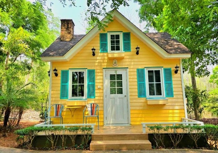 A small yellow house with blue shutters is surrounded by trees.
