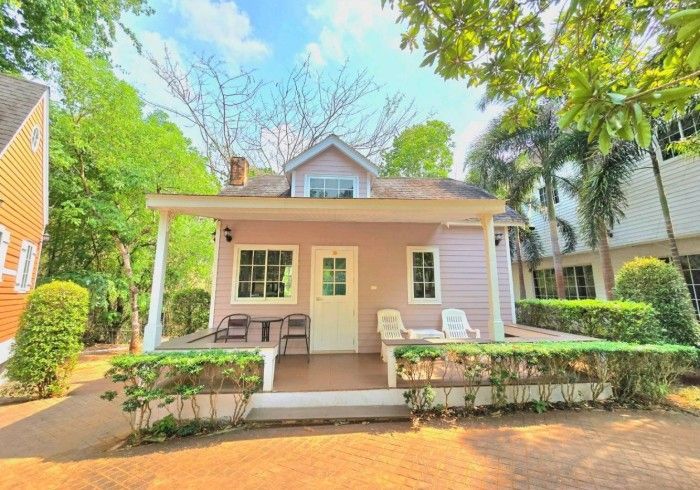 A small pink house with a porch and chairs is surrounded by trees.