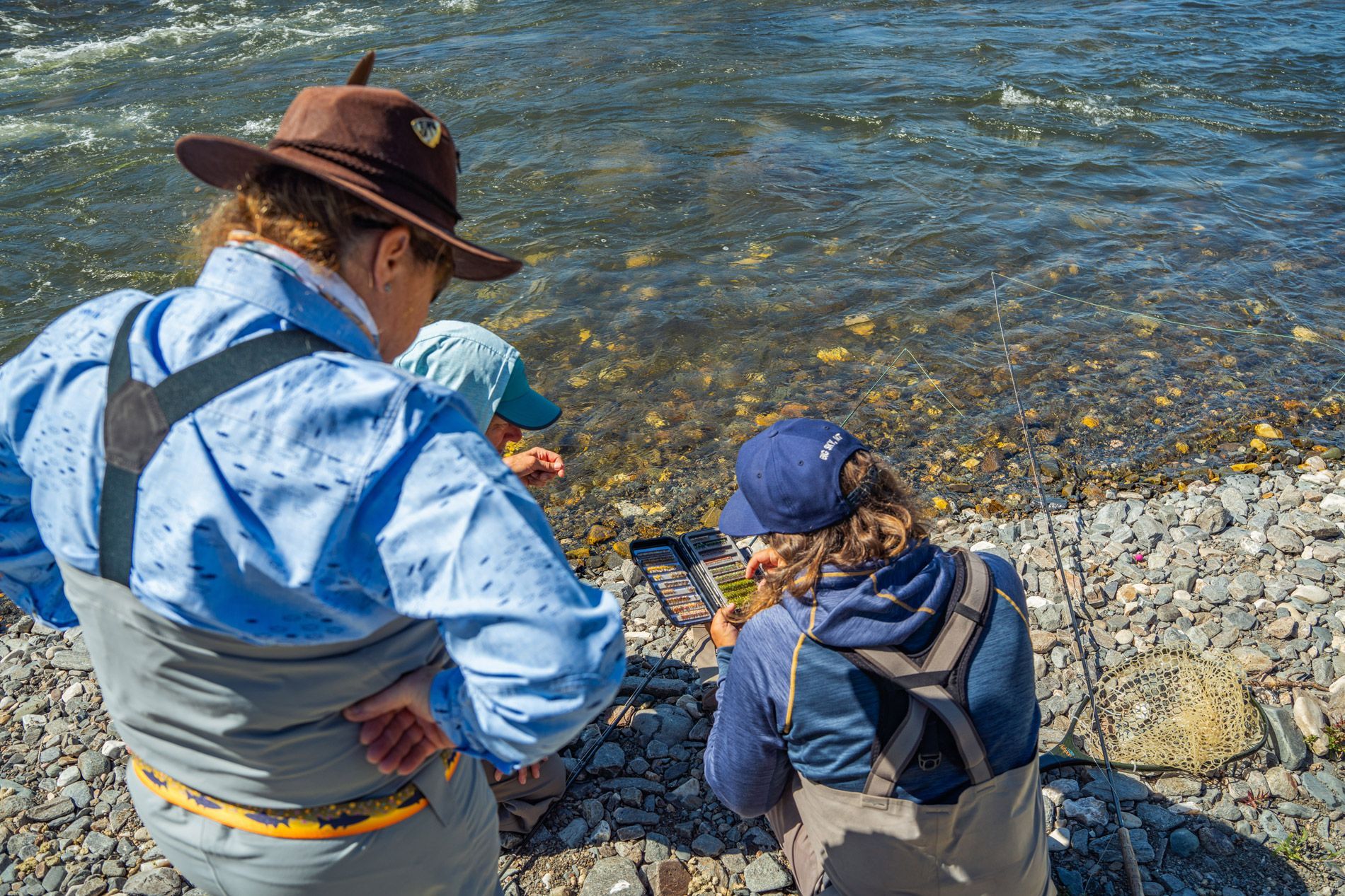 Two people are sitting on the shore of a river looking at a cell phone.