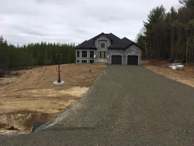 Maison neuve avec allée de gravier, entourée de terre et d'arbres, sous un ciel couvert.