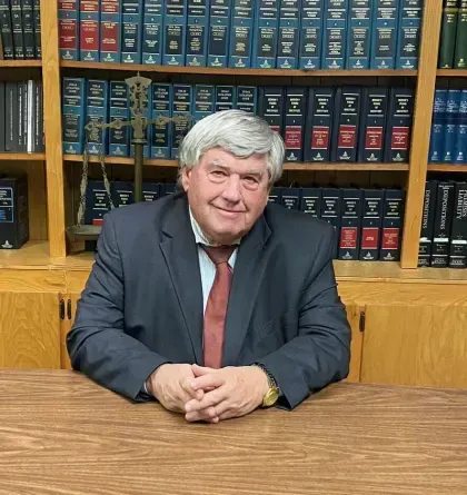 Man in suit at desk, arms crossed, smiling. Bookshelves in background.
